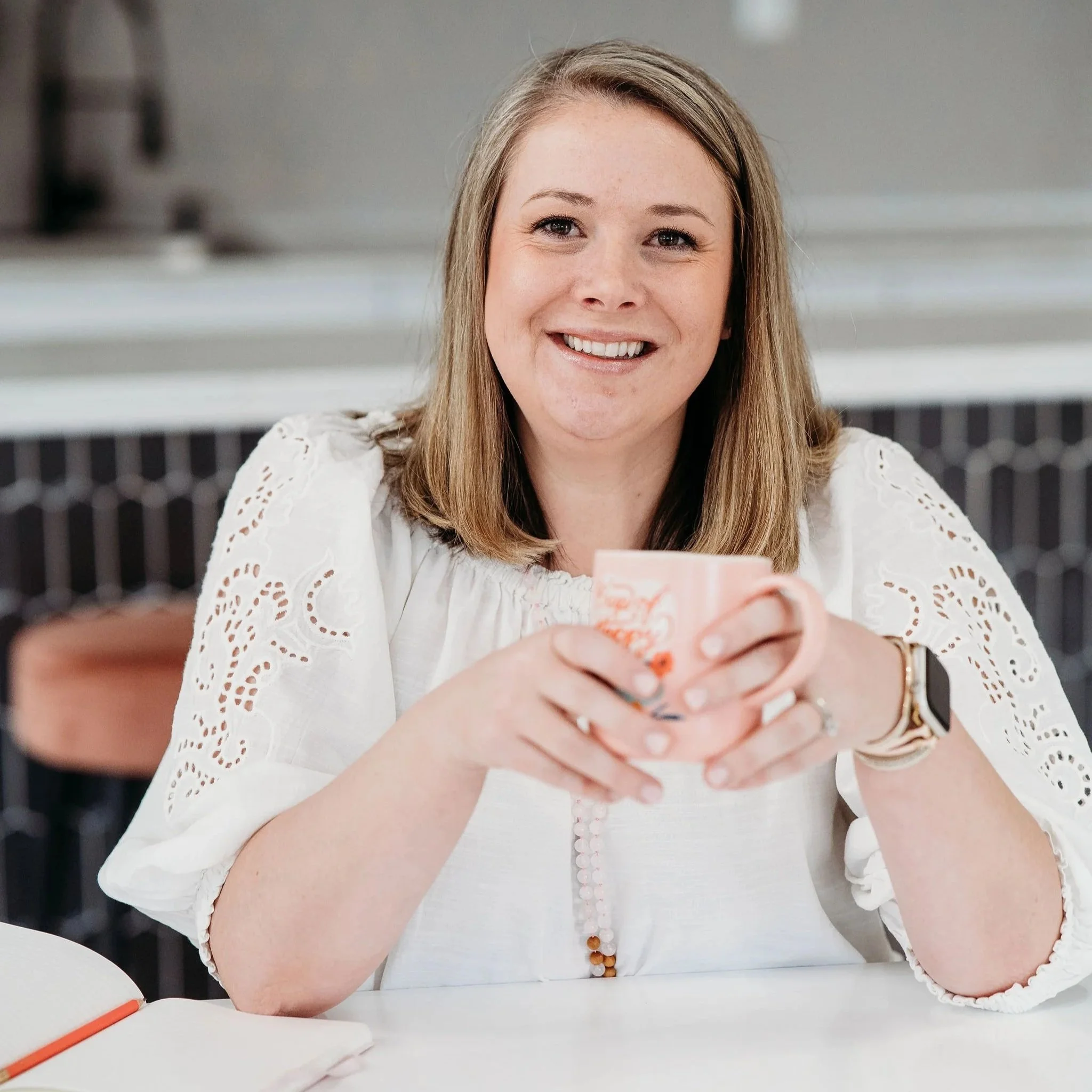 A smiling woman with blonde hair sitting at a white table, holding a paper cup with a floral pattern in front of her, with a blurred background.
