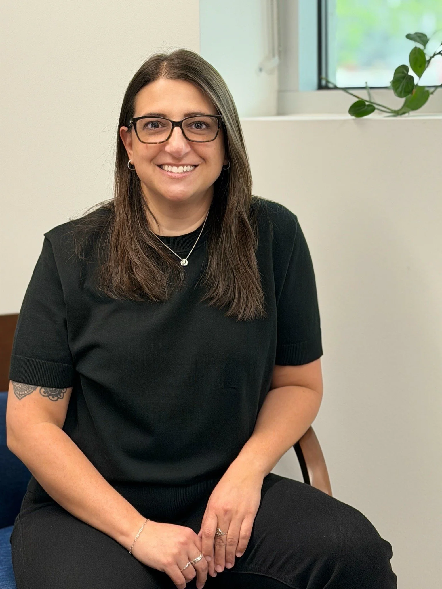 A woman with long brown hair, wearing glasses, a black shirt, and jewelry, sitting in a room near a window with a plant on the windowsill, smiling at the camera.