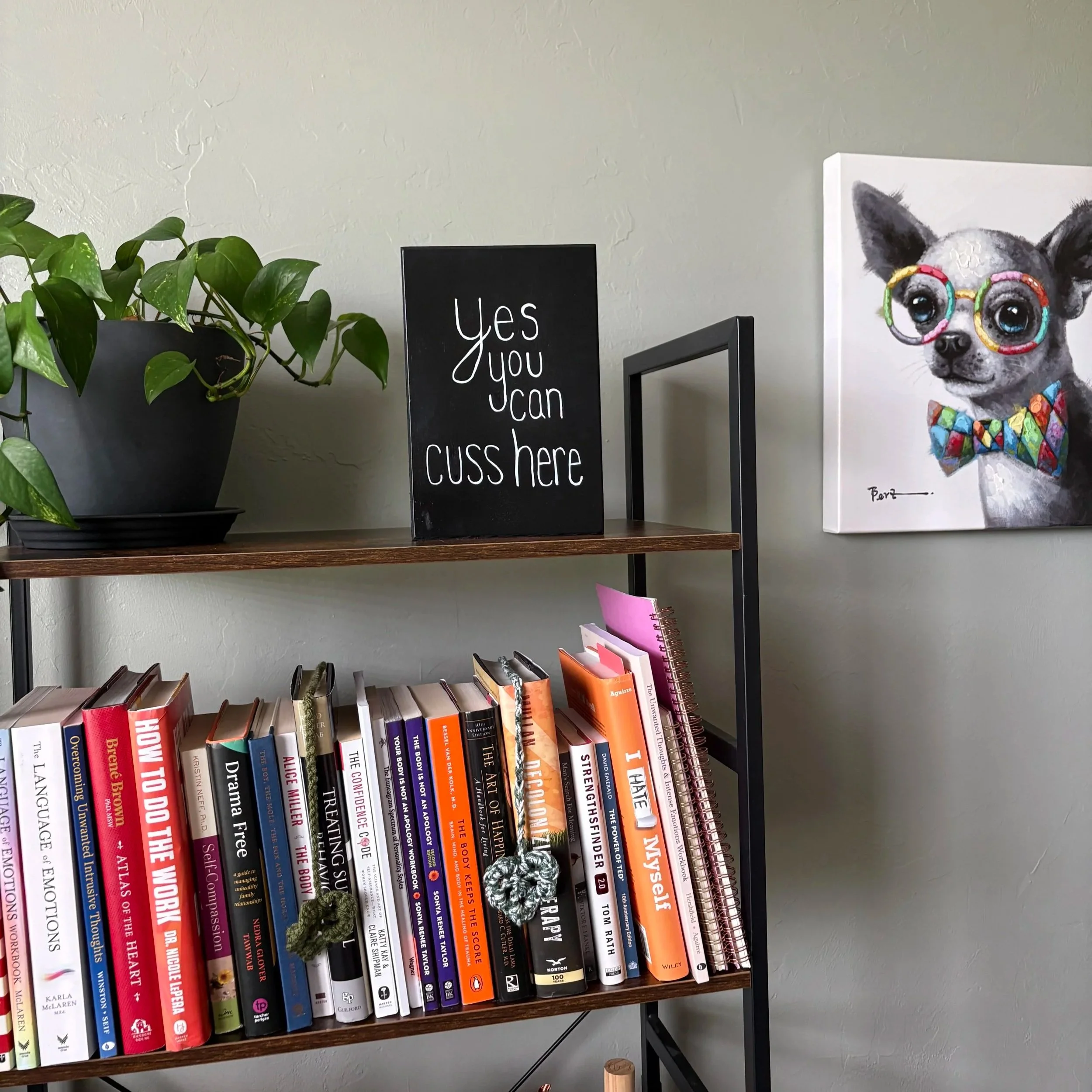 A wooden bookshelf with a row of colorful books on the bottom shelf, a blackboard sign with white writing, a potted plant with green leaves on the left, and a canvas portrait of a dog wearing glasses and a colorful necklace on the right, against a plain light-colored wall.