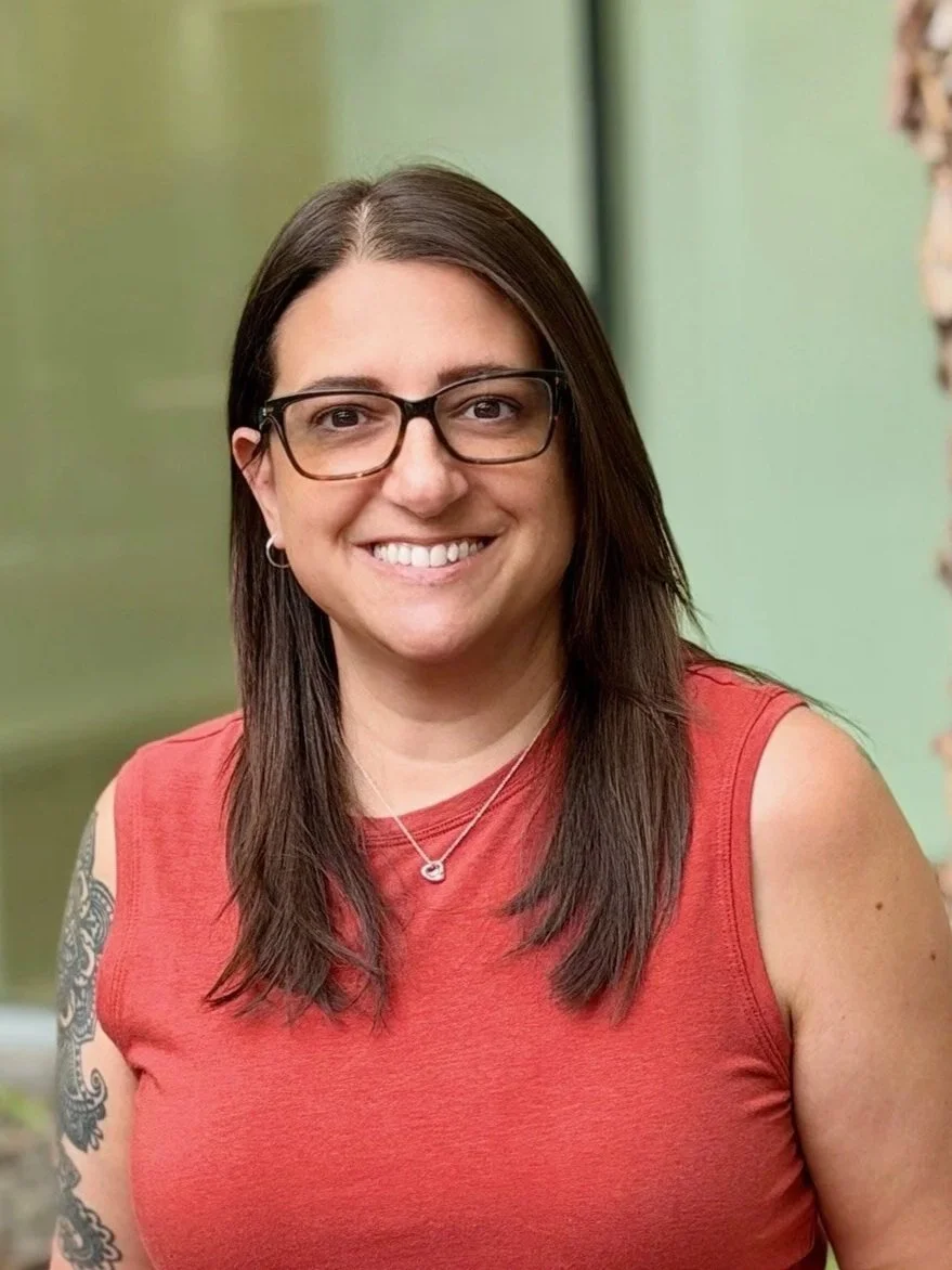A woman with long dark hair, glasses, and earrings, smiling, wearing a sleeveless red top and a necklace, outdoors near a brick wall.