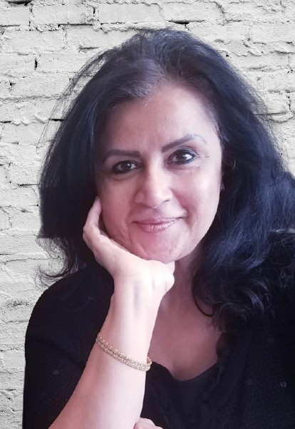 A woman with dark hair, smiling and resting her chin on her hand, standing in front of a brick wall.