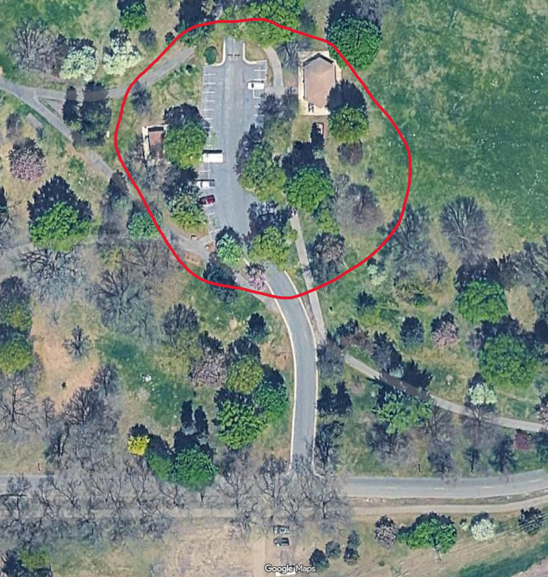 Aerial view of a small park with trees, a parking lot, and a building. The park is surrounded by green areas, roads, and pathways, with a section outlined in red.
