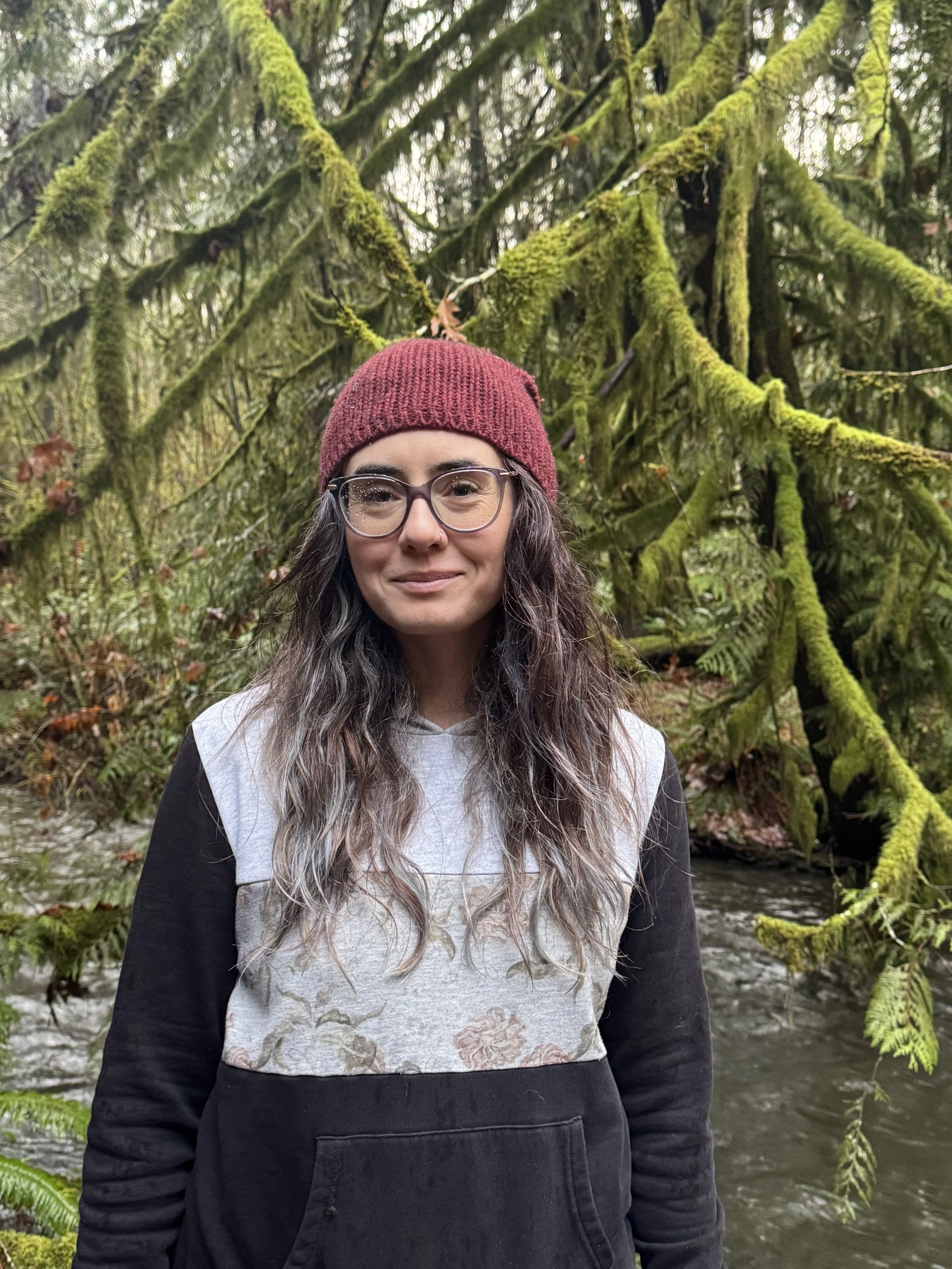 A woman with long wavy hair, glasses, and a red knit beanie, standing outdoors near a moss-covered tree and a small stream.
