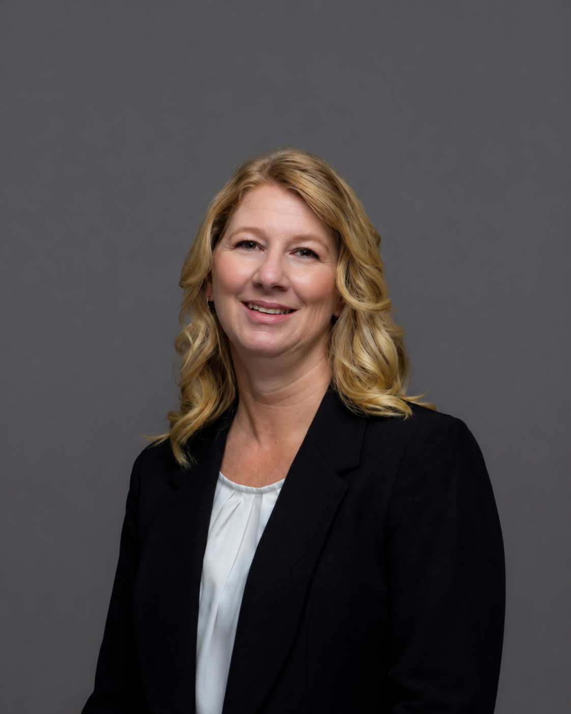A professional woman with wavy blonde hair, wearing a black blazer over a white top, smiling against a plain gray background.