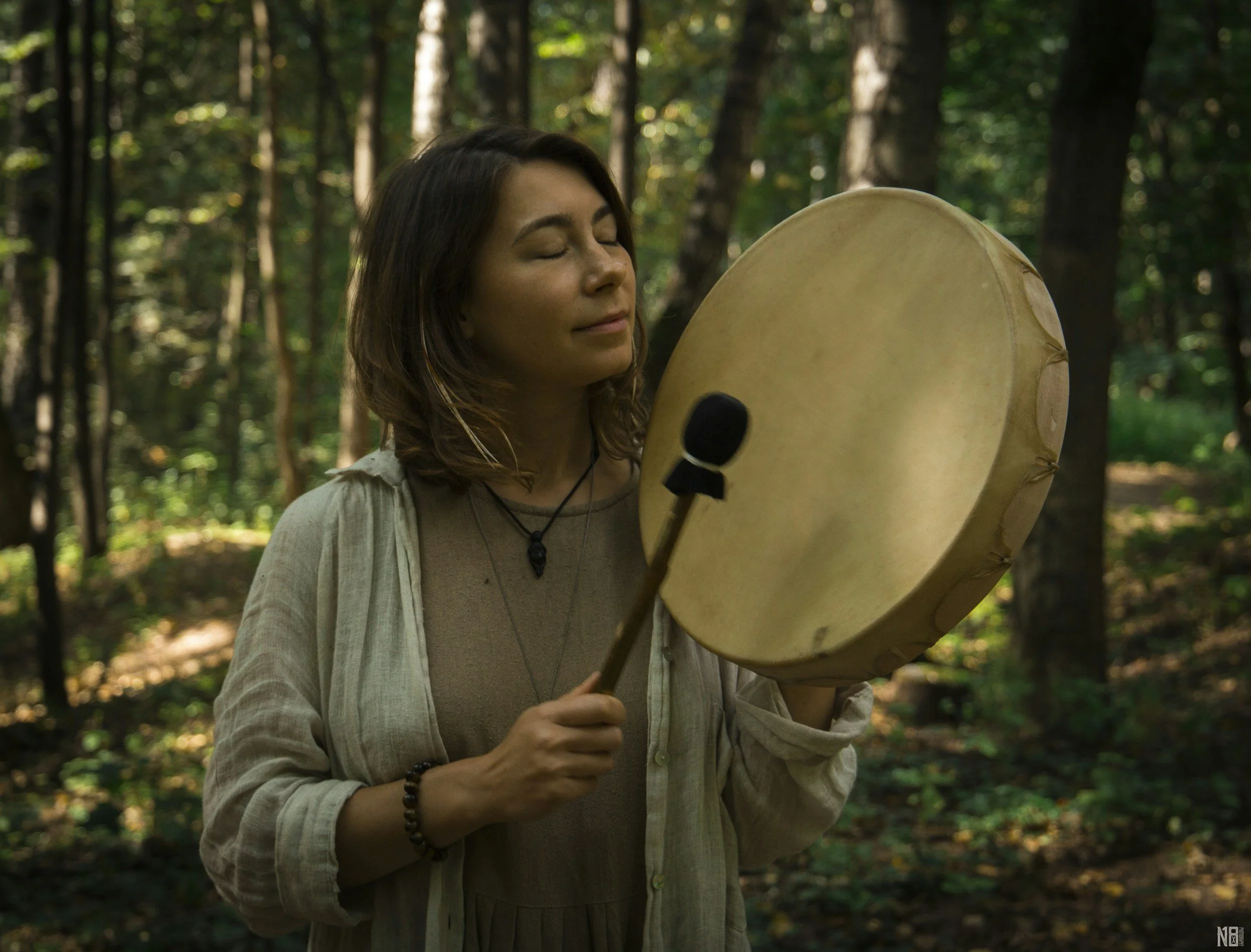 A woman with dark hair, wearing a beige shirt and jewelry, holds a drumstick in her right hand and plays a large hand drum outdoors in a forest with greenery and trees in the background.