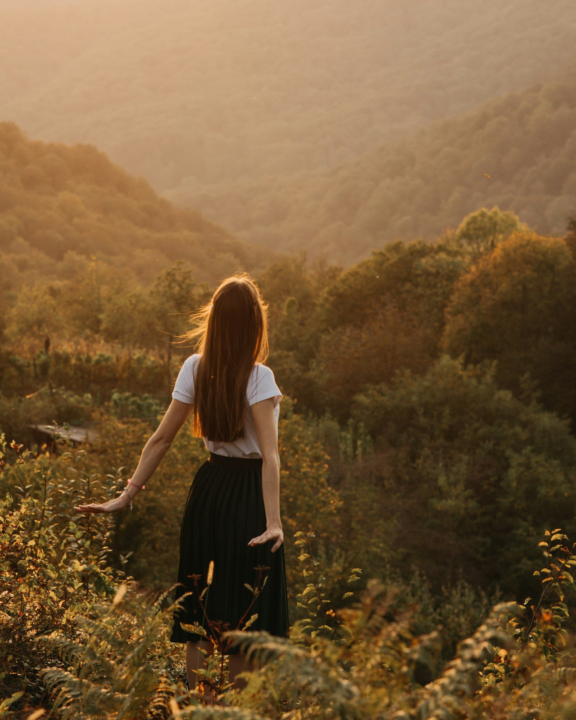 A woman with long hair, wearing a white top and a black skirt, standing in a field of wildflowers with a forested hillside and mountains in the background during sunset.
