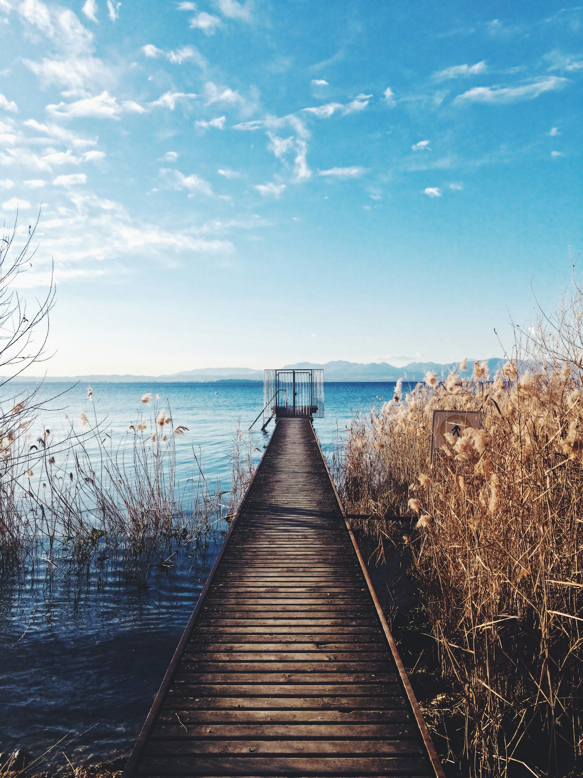 Wooden dock extends over a body of water with dry grass on either side, leading to a small viewing platform against a backdrop of distant mountains and a partly cloudy sky.