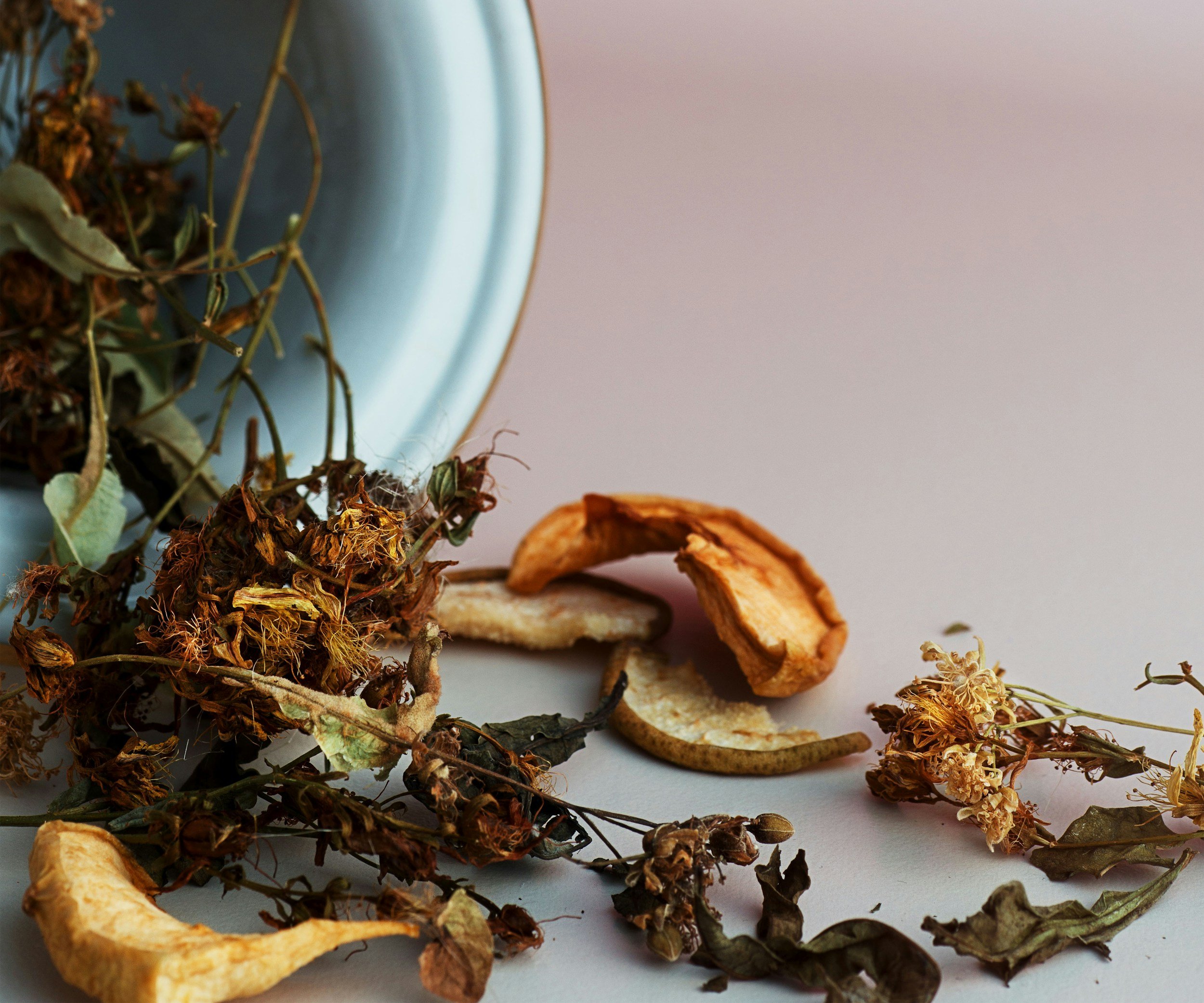 Various dried herbs and flower petals scattered on a white surface, with a light blue vase partly visible in the background.