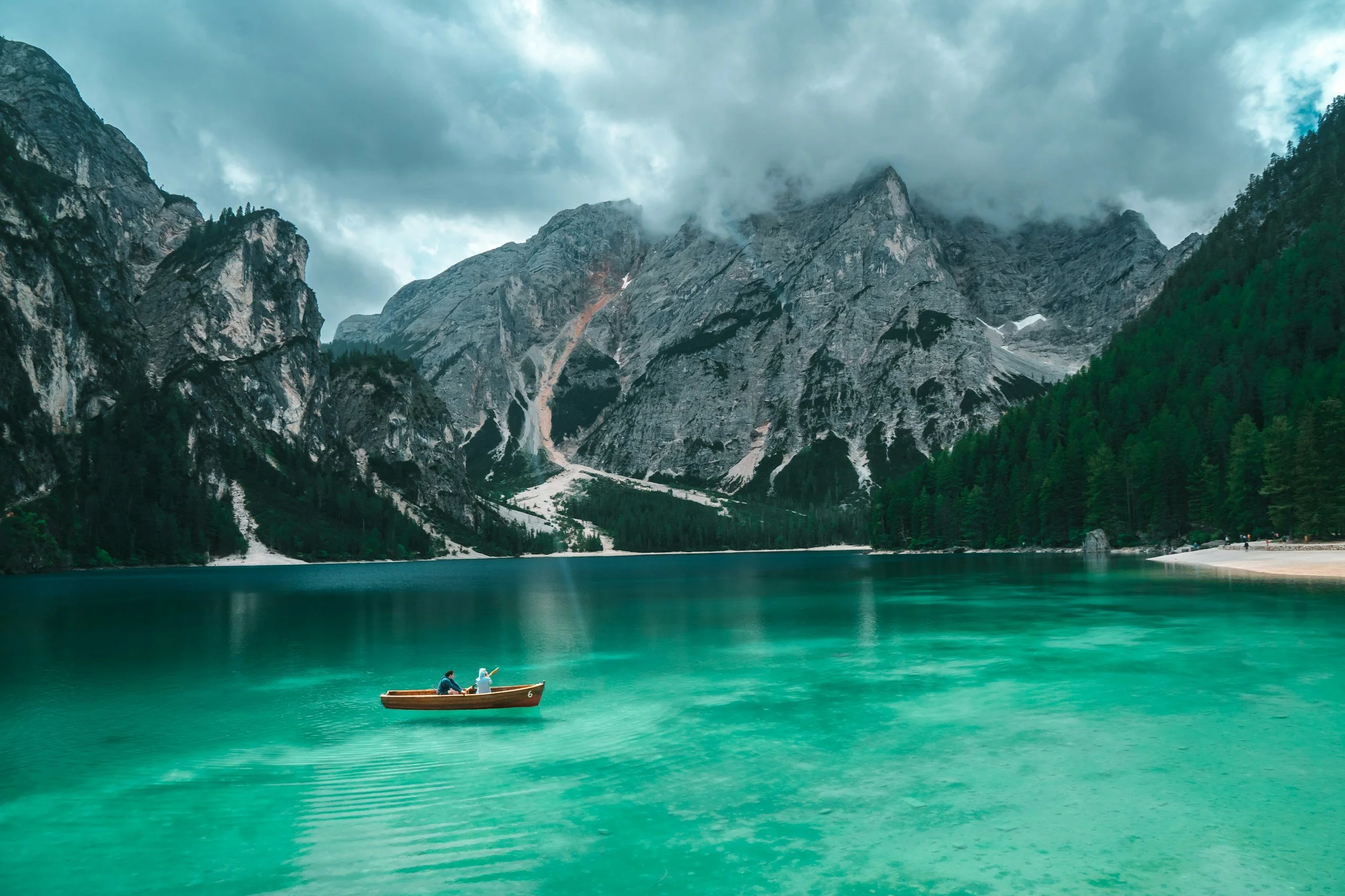 A small wooden boat with two people on a turquoise lake surrounded by towering mountains with patches of snow and forested slopes, under a cloudy sky.