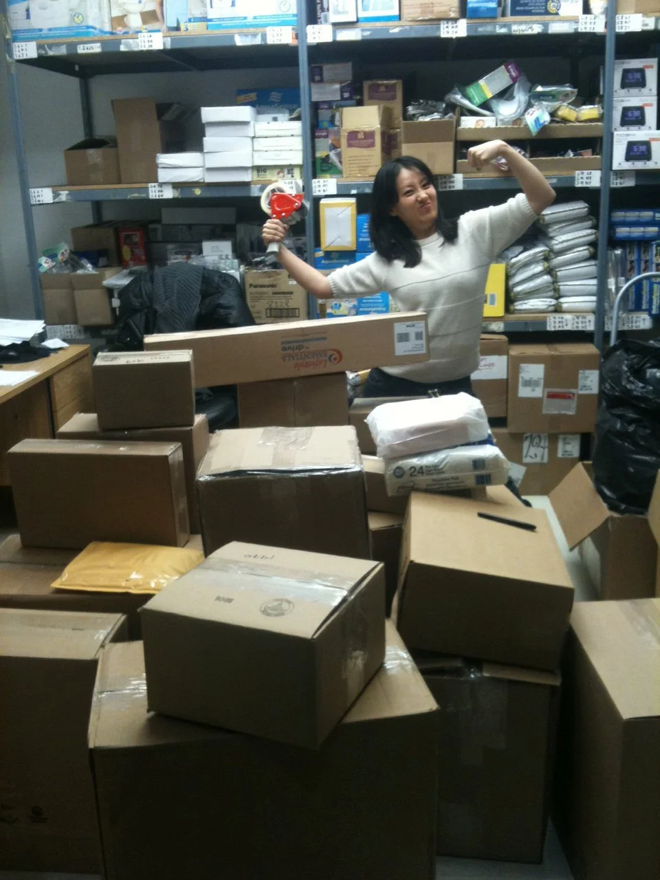 Woman in a warehouse with cardboard boxes, holding a red and white kettle, flexing her arm, smiling.