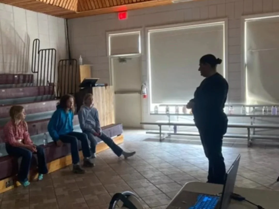 A woman is standing and speaking in front of three children who are seated on bench seats in a room with wooden walls and a tiled floor. There is a laptop on a table in the foreground and a fire extinguisher near the windows.