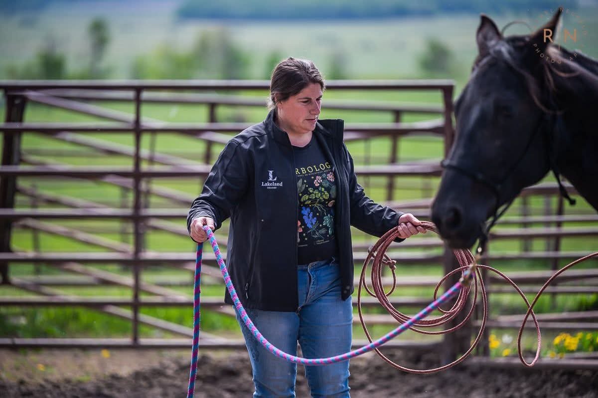 A woman with dark hair, wearing a black jacket and jeans, holds a colorful rope and a coiled lasso in a fenced outdoor area with a horse on the right side of the image.