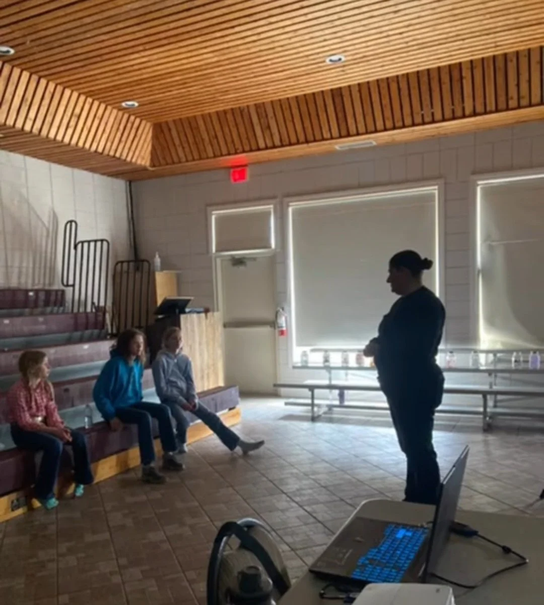 A woman standing in front of three seated children in a room with wooden ceiling and tiled floor, with a laptop on a table in the foreground.