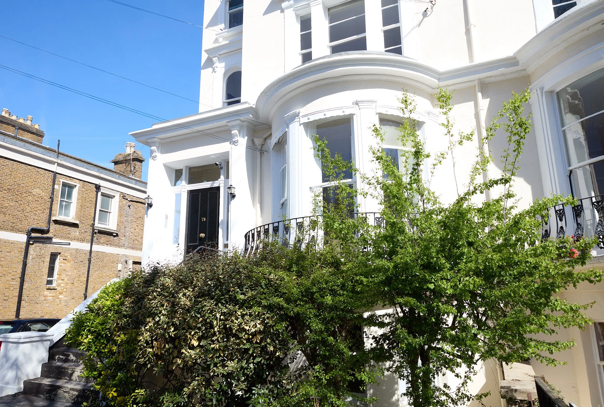 White multi-story house with bay windows, black front door, elaborate architectural details, surrounded by green shrubs and a tree, with neighboring brick building and power lines in the sky.