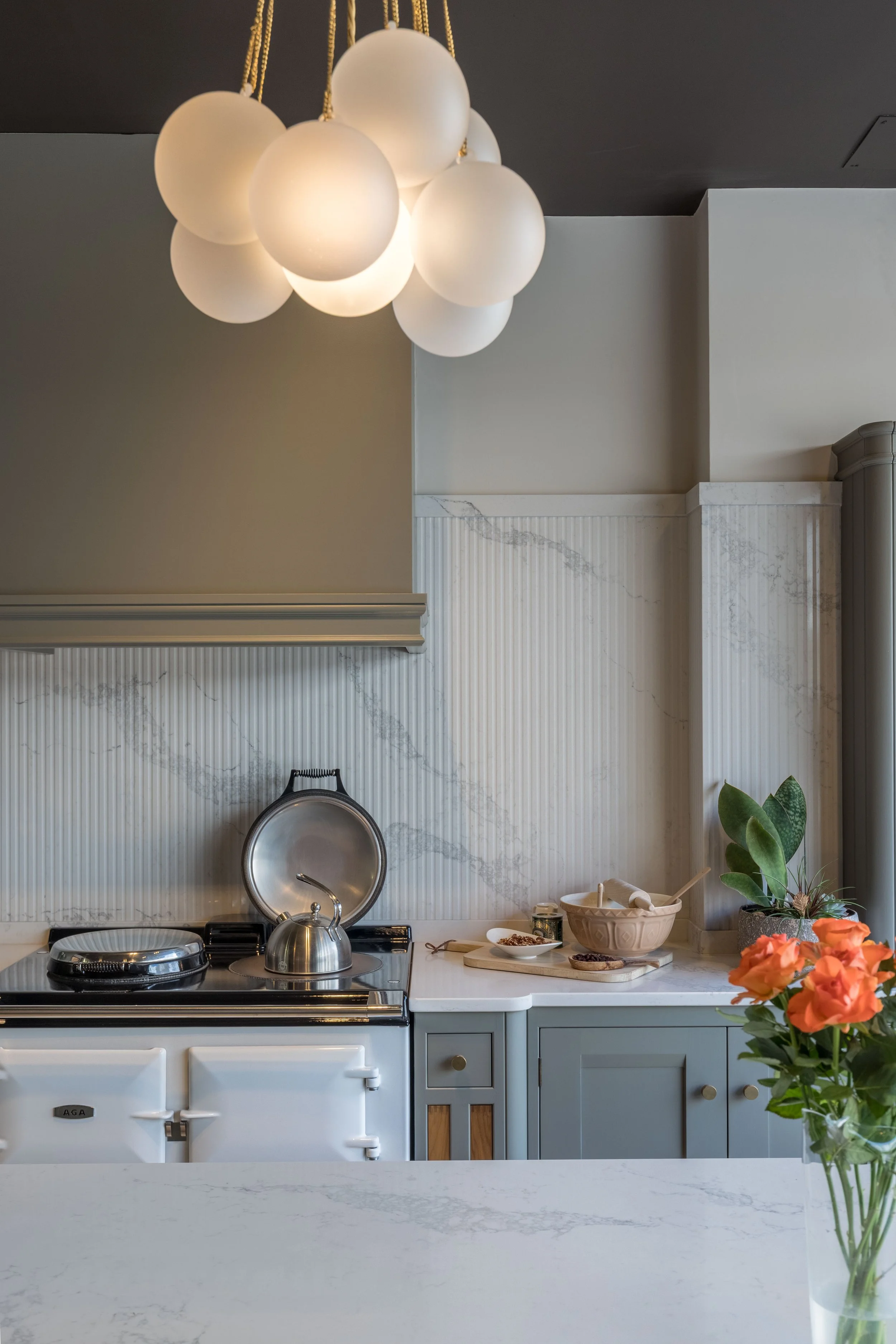 Modern kitchen with a white stove, stainless steel kettle, beige marble backsplash, light blue cabinets, and a vase of orange roses on the white marble countertop. A pendant light fixture with round frosted glass globes hangs from the dark ceiling.
