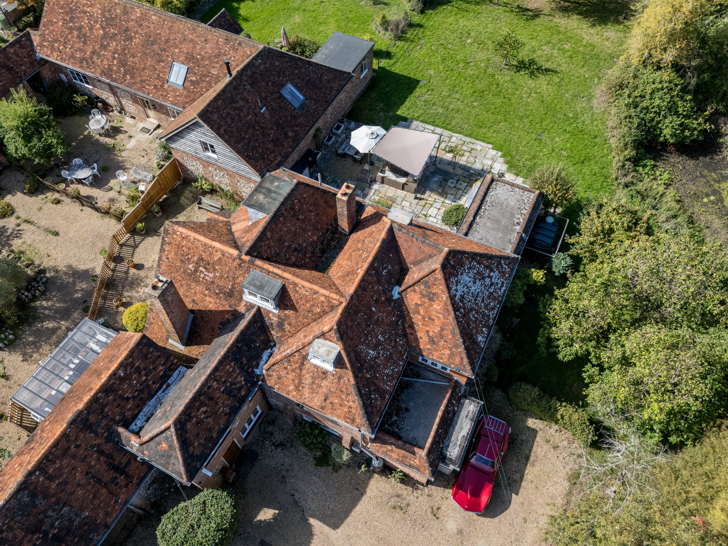 Aerial view of a house with a red brick roof, outdoor furniture with a white umbrella on a patio, green lawn, and surrounding trees.