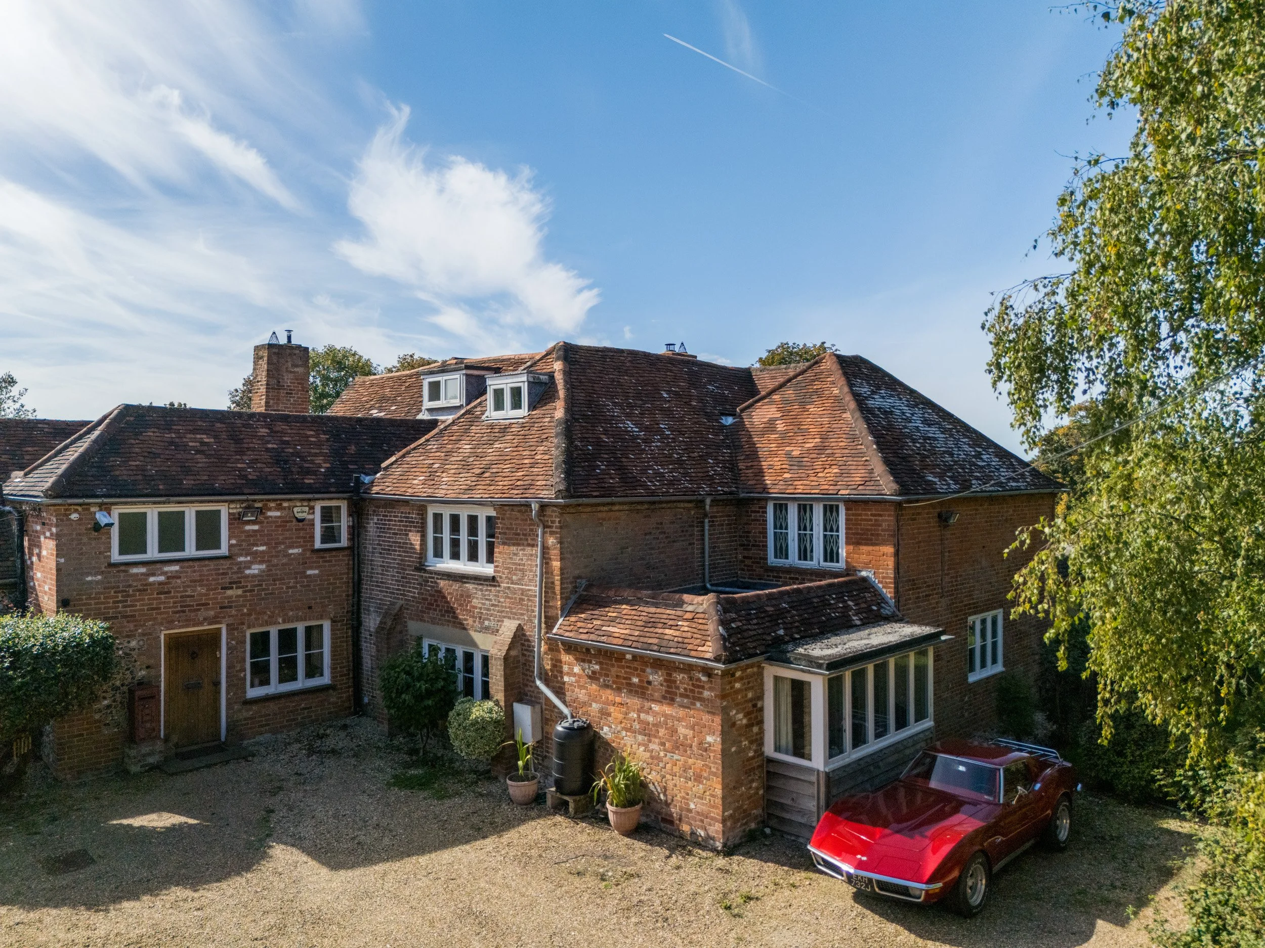 The image shows the roofs of two brick houses with terracotta tiles, a chimney, two small dormer windows on the roof of one house, and a large window on the ground floor. There is a clear blue sky with some clouds and an airplane contrail. On the rig