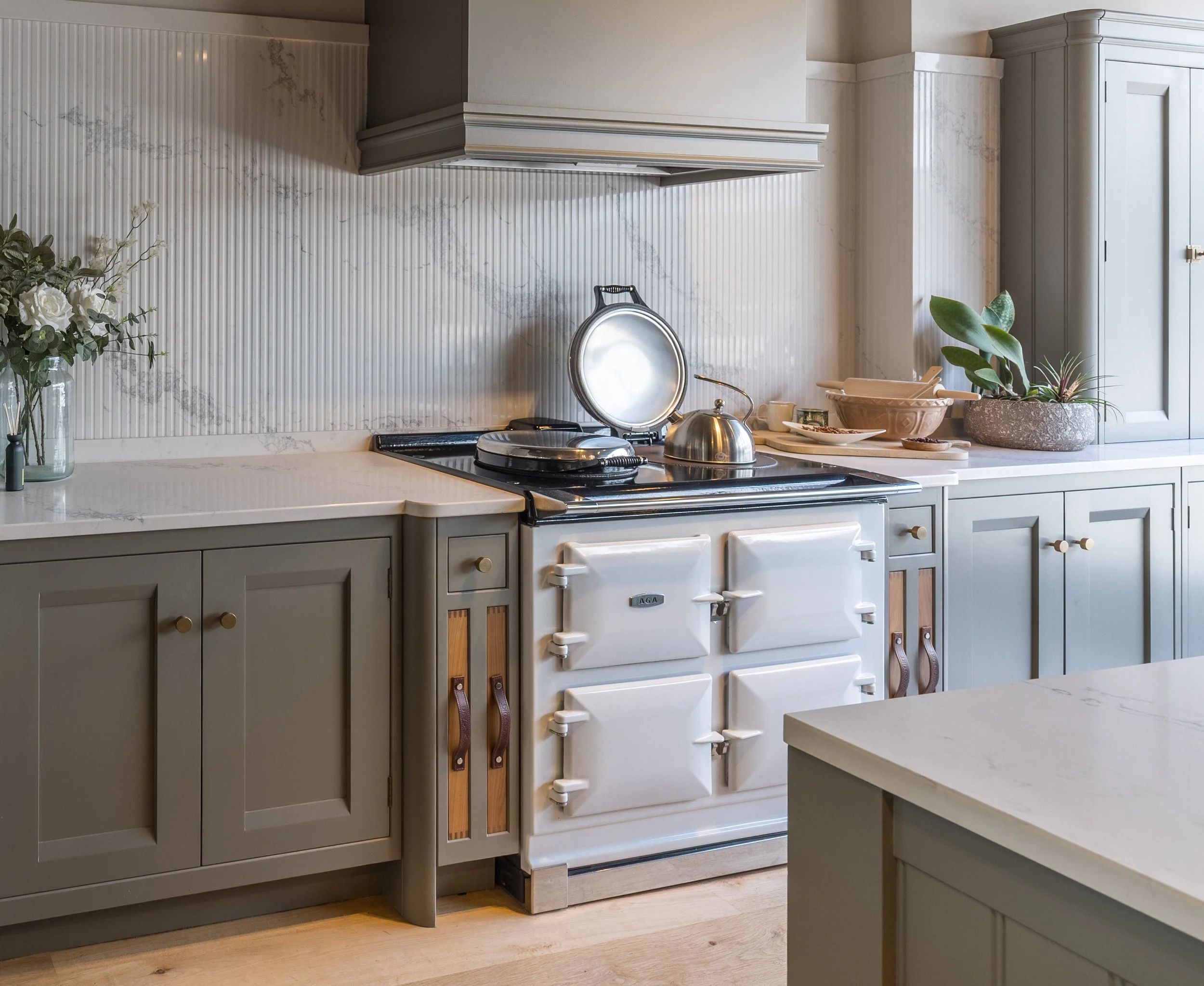A kitchen with light grey cabinets, a white marble countertop, and a backsplash with vertical beadboard paneling and marble accents. On the counter, there is a silver kettle, a silver pot, and a black burner. To the left, a glass vase with white roses and greenery. On the right, a large round planter with a green plant, and a beige bowl with utensils.