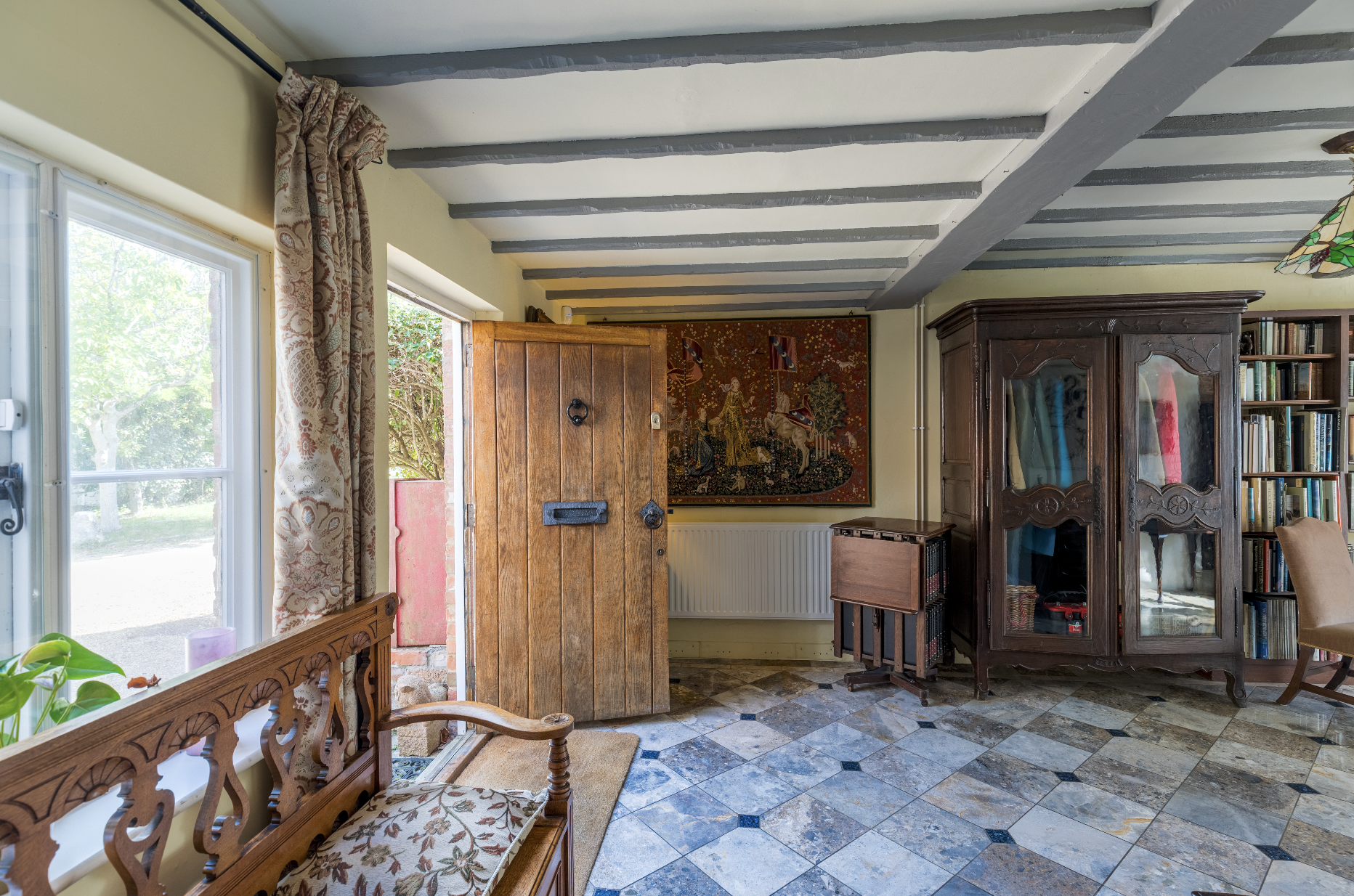Interior view of a cozy living room showing a wooden door, a large ornate vintage cabinet, a bookshelf, a tapestry on the wall, and a window with floral curtains, sunlight coming through.
