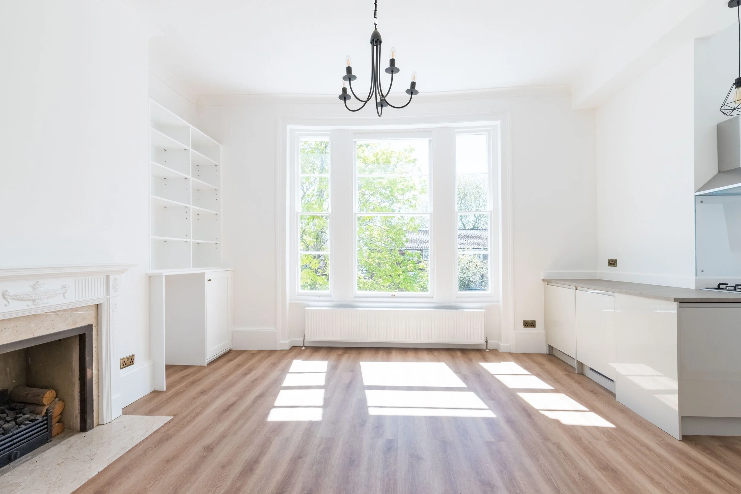 Bright kitchen with large window, white cabinets, and a chandelier, featuring hardwood floors and a fireplace with a decorative mantle.