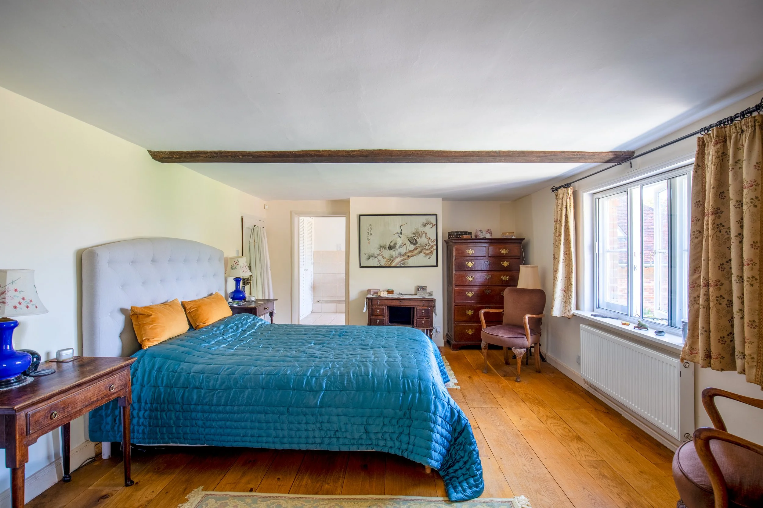 A bedroom with a bed featuring a tufted white headboard, blue quilt, and orange pillows. There are two wooden nightstands with blue lamps on each side of the bed. The room has hardwood flooring and is decorated with curtains, artwork, and antique fur