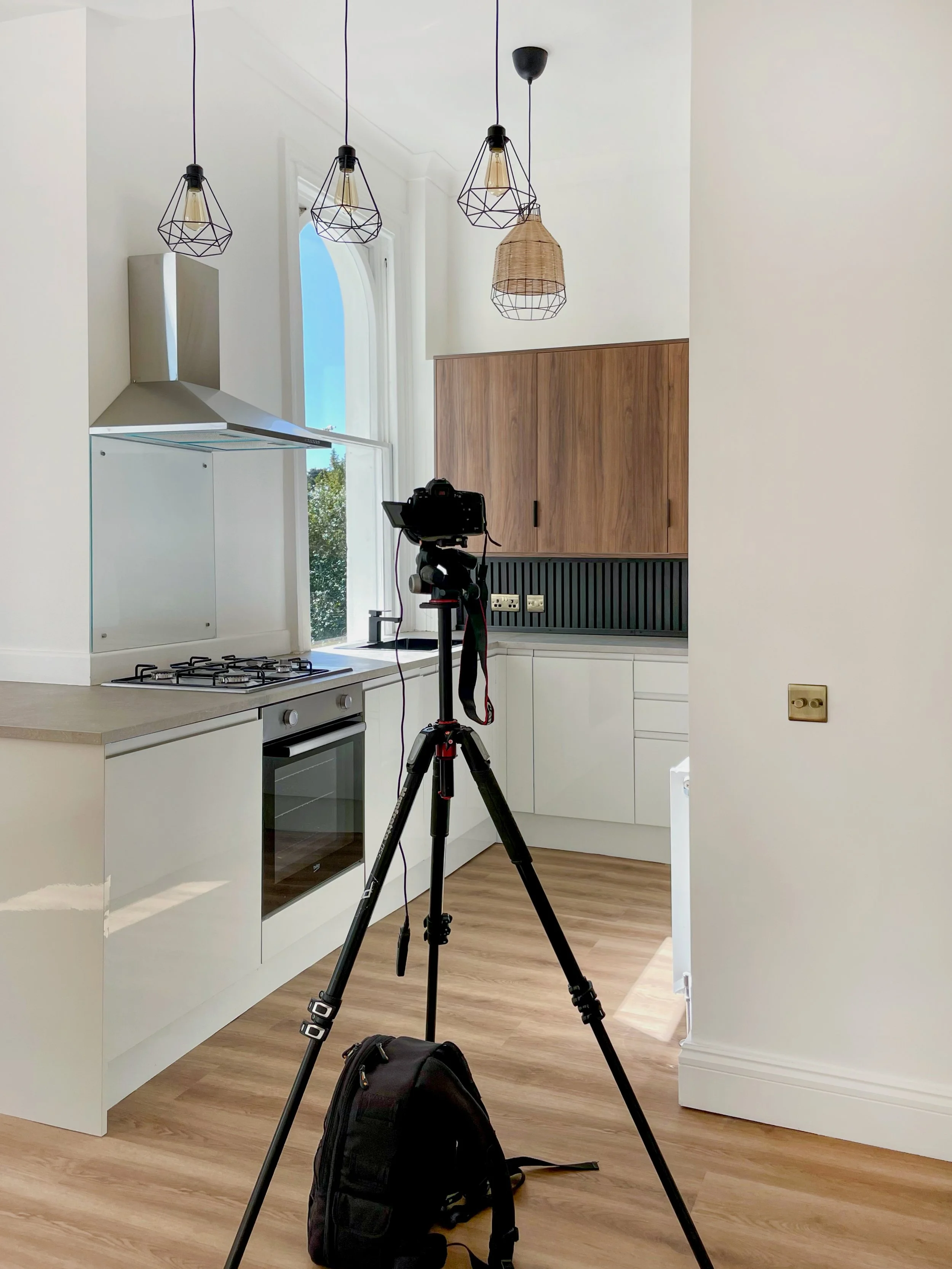 A kitchen with white cabinets and wooden flooring, a tripod with a camera set up facing the counter, and a black backpack on the floor. Hanging pendant lights and a window with blue sky visible outside.