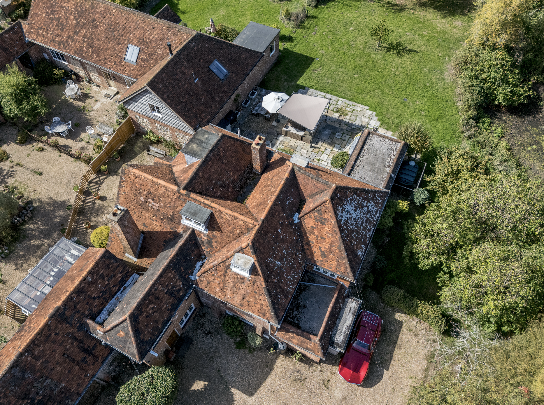 Aerial view of a residential backyard showing multiple red-tiled roofs of brick houses, green lawn, patio with umbrellas and outdoor furniture, a red vehicle parked nearby, and various trees and shrubs surrounding the area.