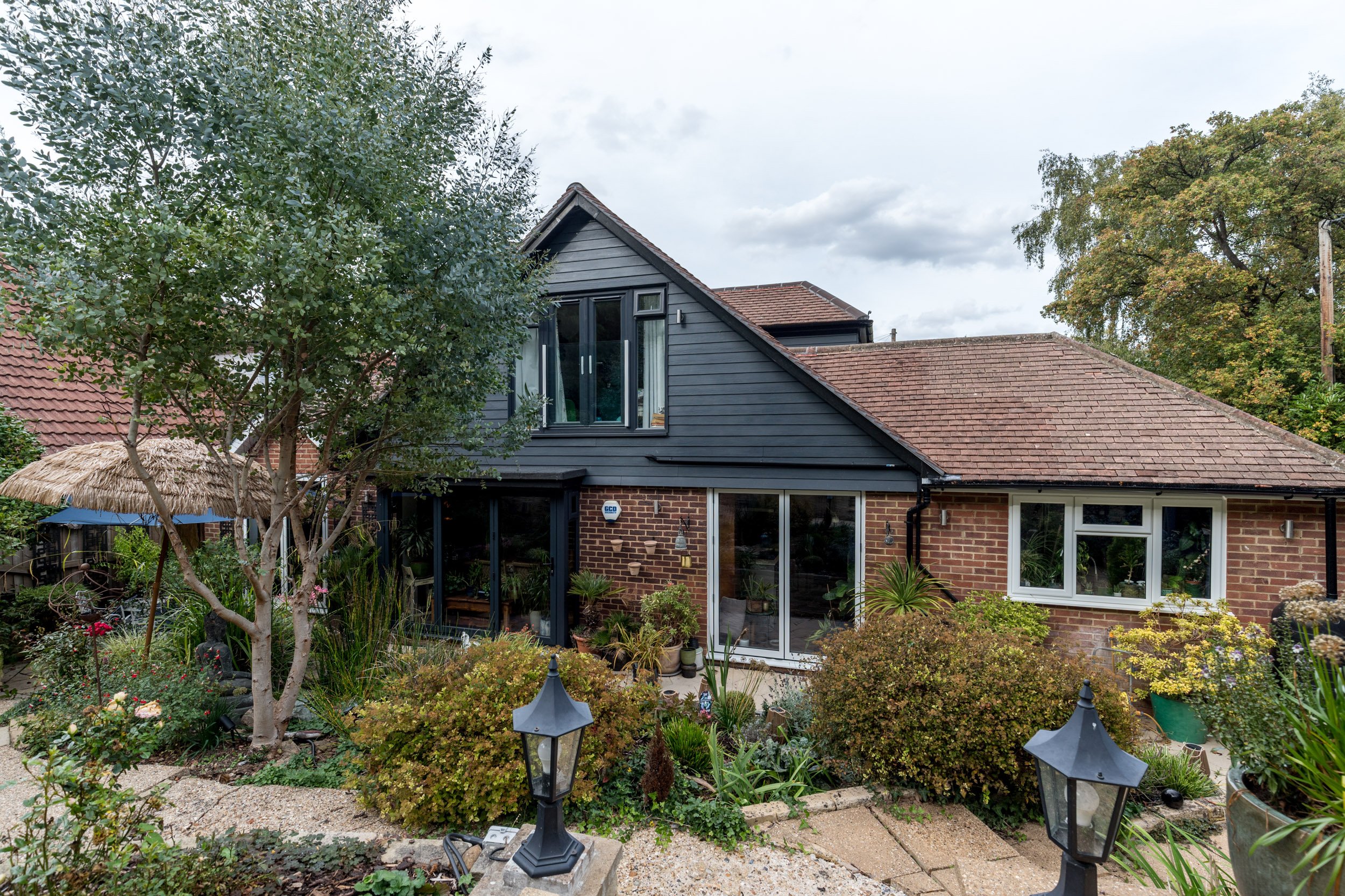 View of a house with grey and brick exterior, garden with plants and pathway, trees, and outdoor lanterns.
