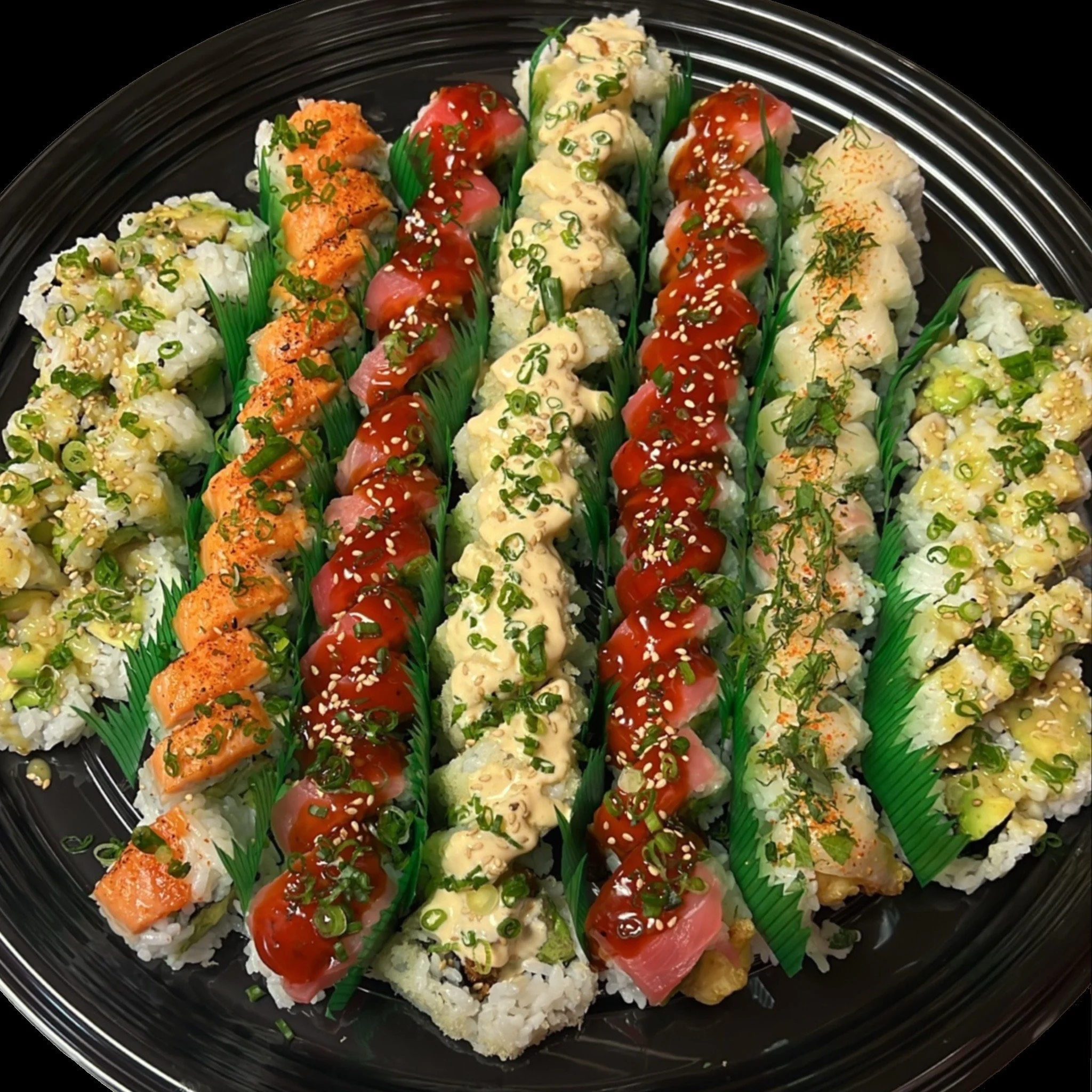 Assorted sushi rolls arranged on a black plate, garnished with chopped green onions, sesame seeds, and decorative plastic grass.
