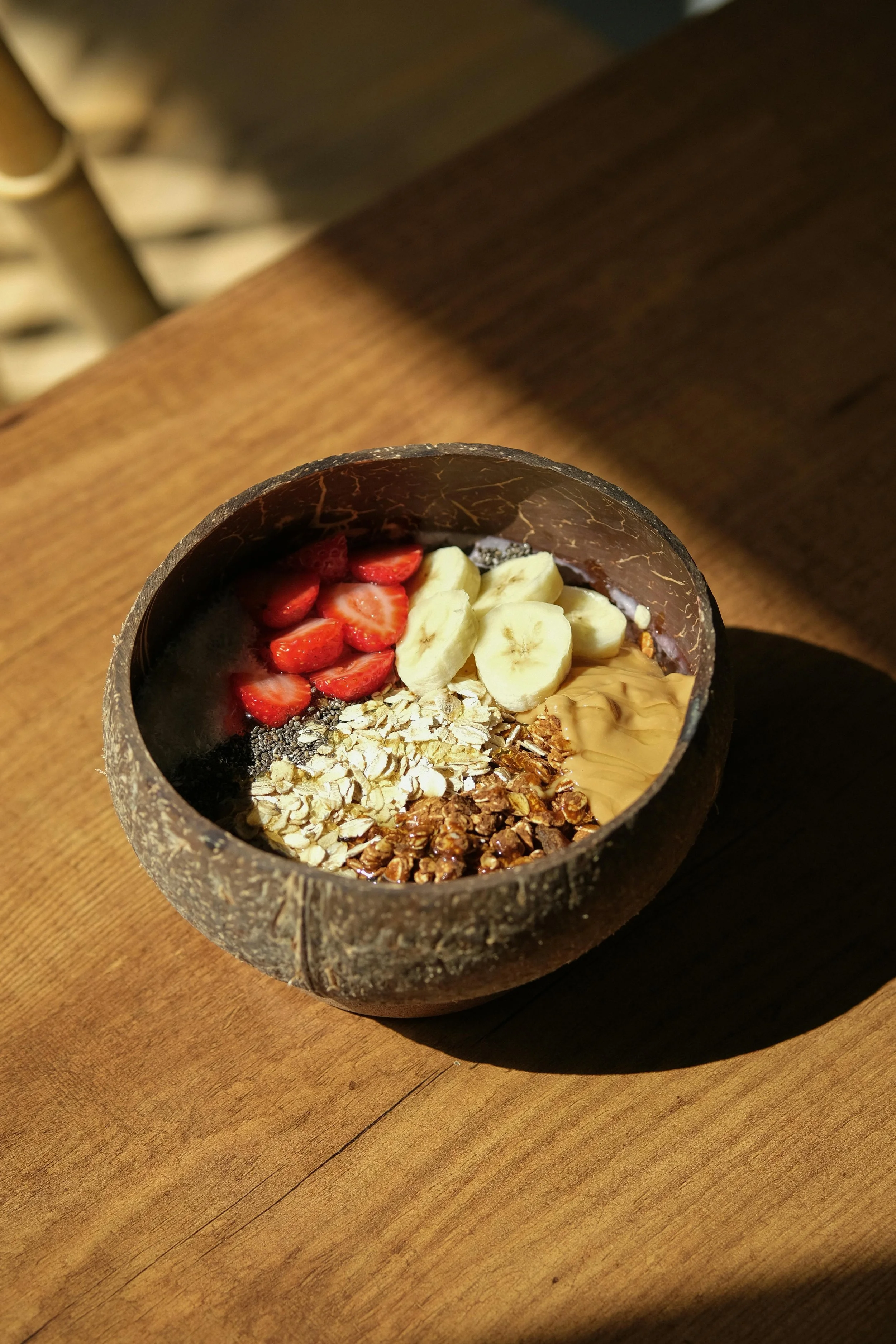 A bowl filled with sliced strawberries, banana pieces, granola, chia seeds, and peanut butter on a wooden table.