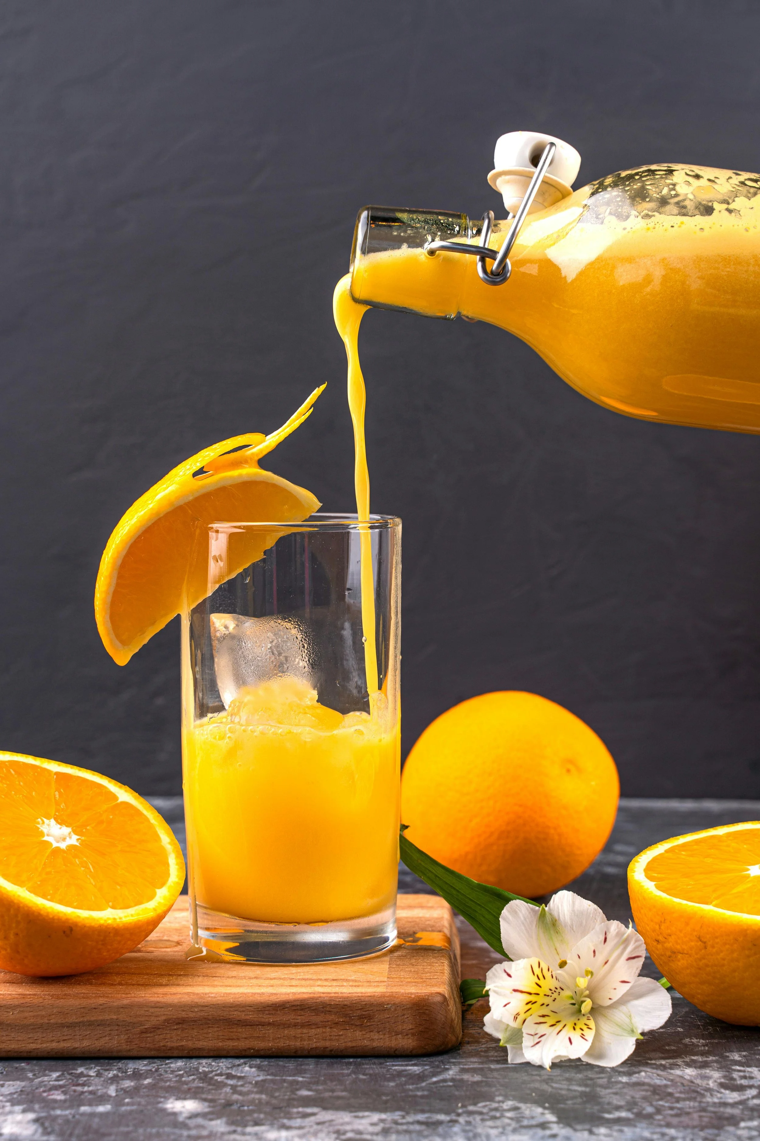 A glass of orange juice with an orange slice on the rim, pouring from a glass bottle into the glass, surrounded by whole and halved oranges, laid on a wooden board on a gray surface with a gray background and white flowers.