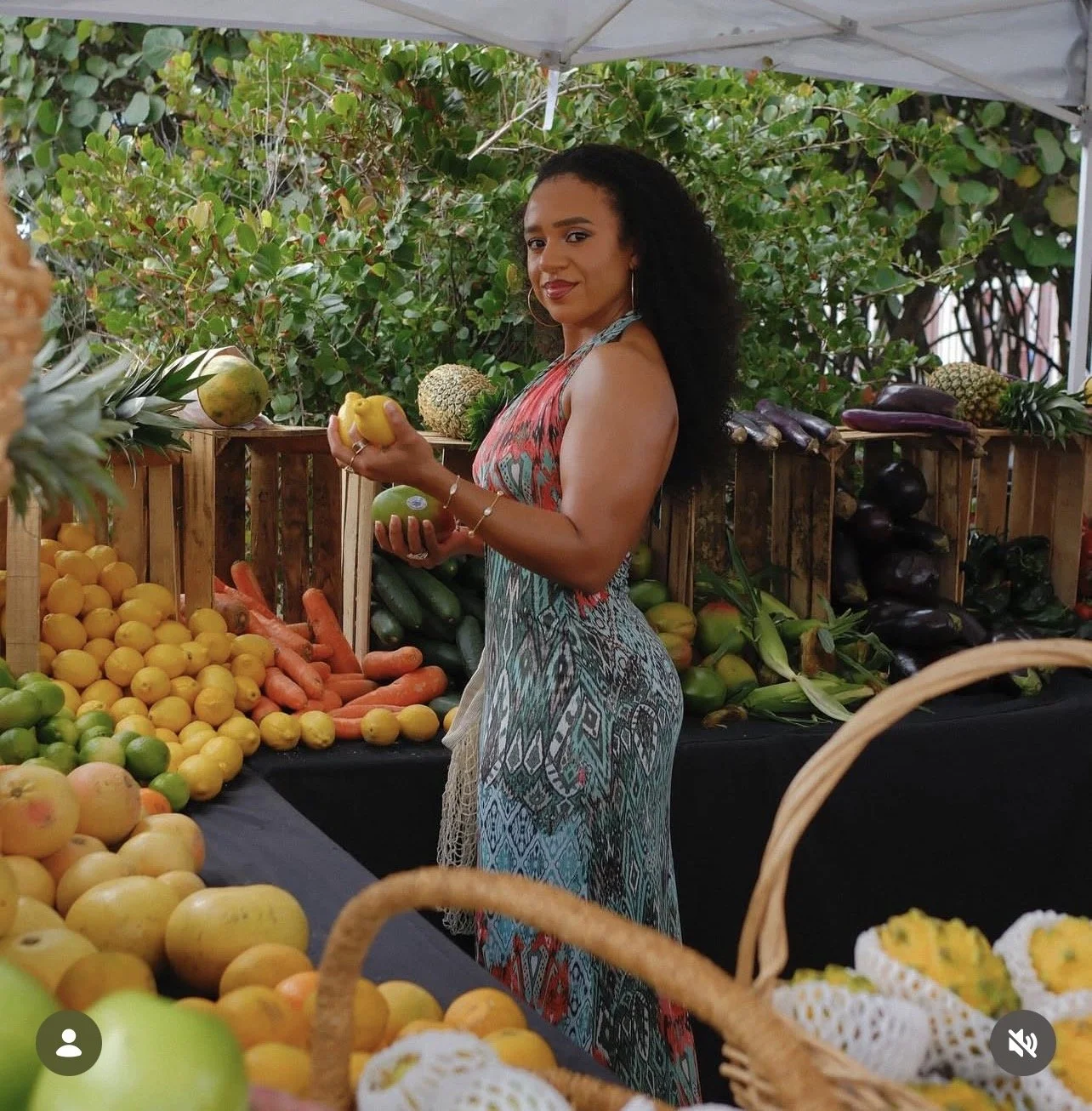 A woman shopping for fresh fruits and vegetables at an outdoor market, holding a yellow fruit and surrounded by crates of produce such as carrots, lemons, and zucchini.