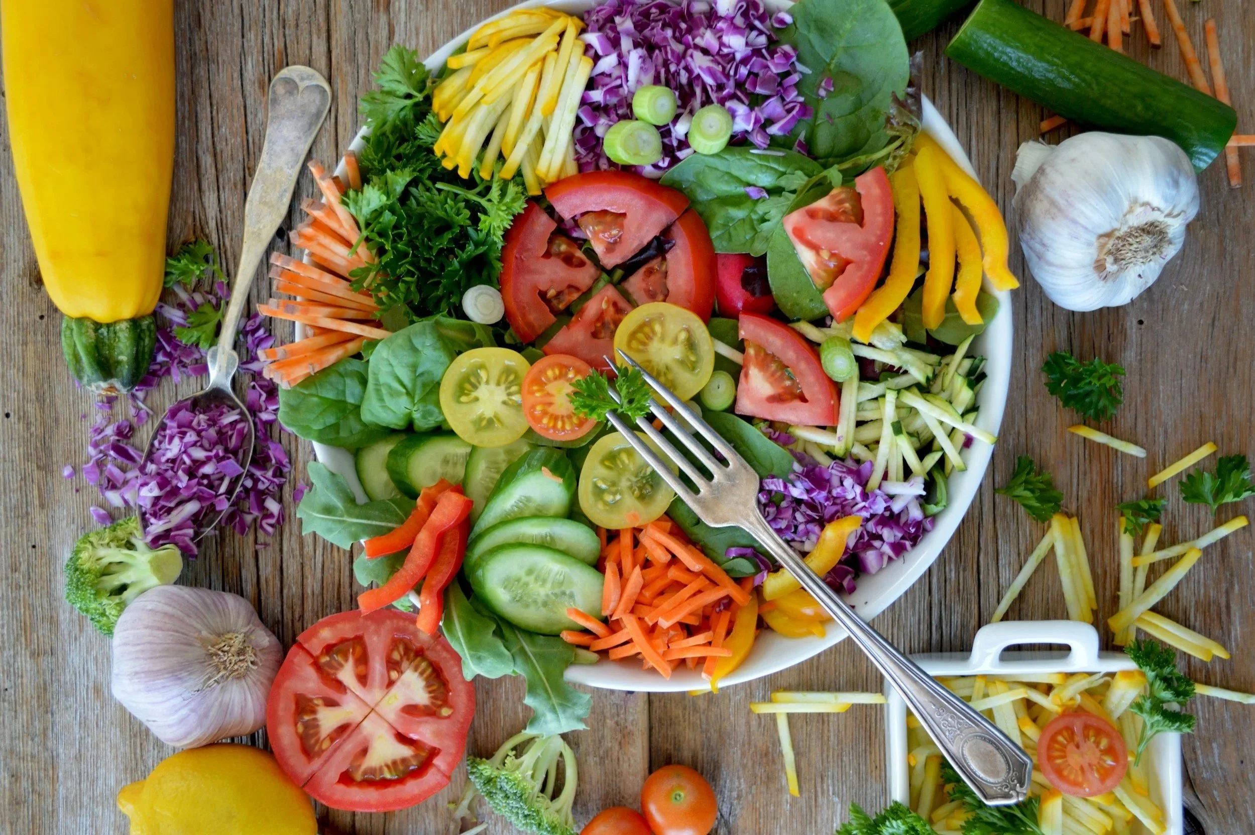 A colorful salad on a wooden table, containing sliced tomatoes, cucumbers, shredded cheese, shredded carrots, purple cabbage, yellow bell peppers, green spinach leaves, scallions, parsley, and garnished with lemon and garlic.