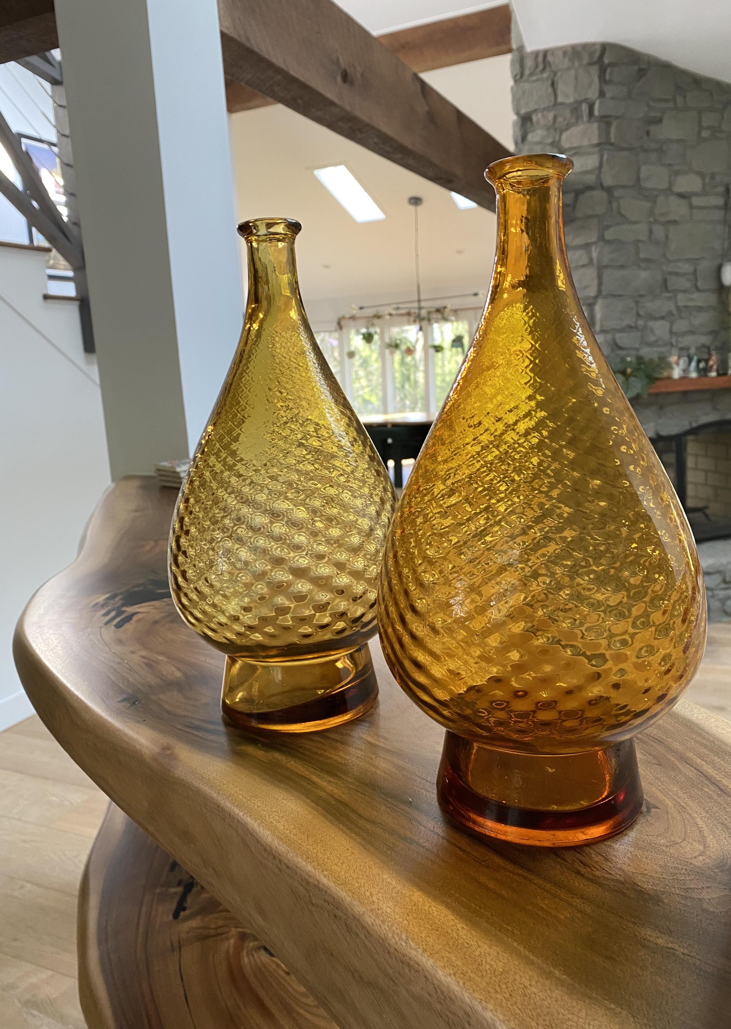 Two amber-colored glass vases with textured patterns placed on floating walnut shelves in a living room setting with a stone fireplace and large windows in the background.