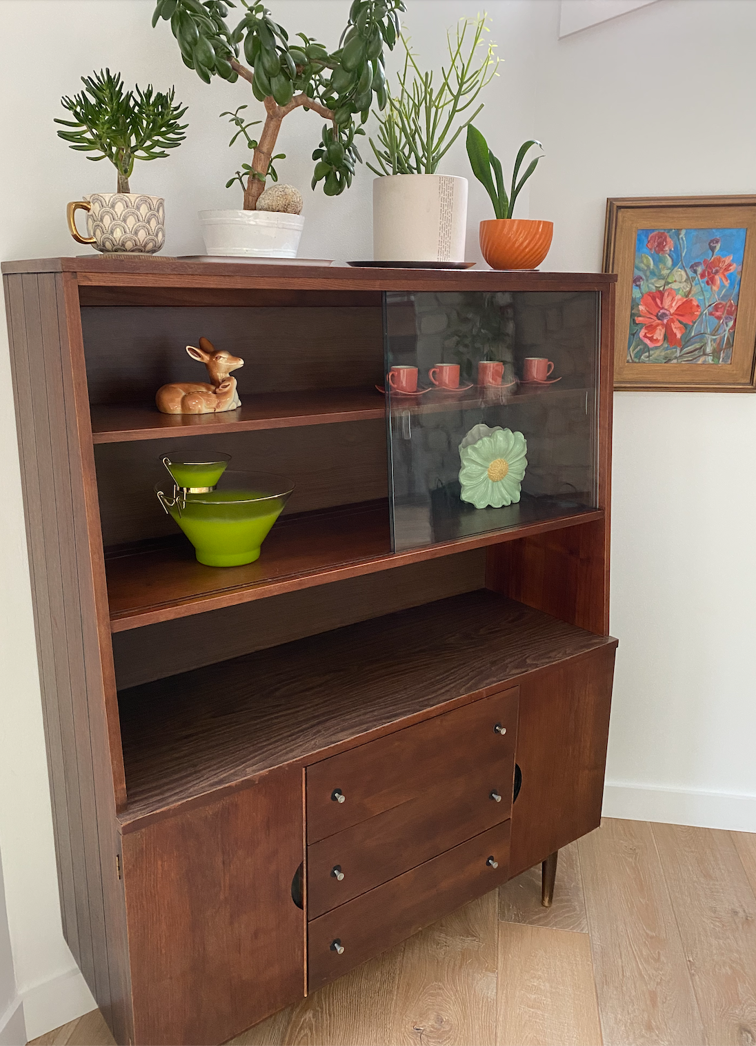 A mid-century wooden display cabinet with glass doors, decorated with potted plants on top, a framed floral painting on the wall, and decorative items inside including ceramic cups, a deer figurine, green bowls, and a ceramic flower.