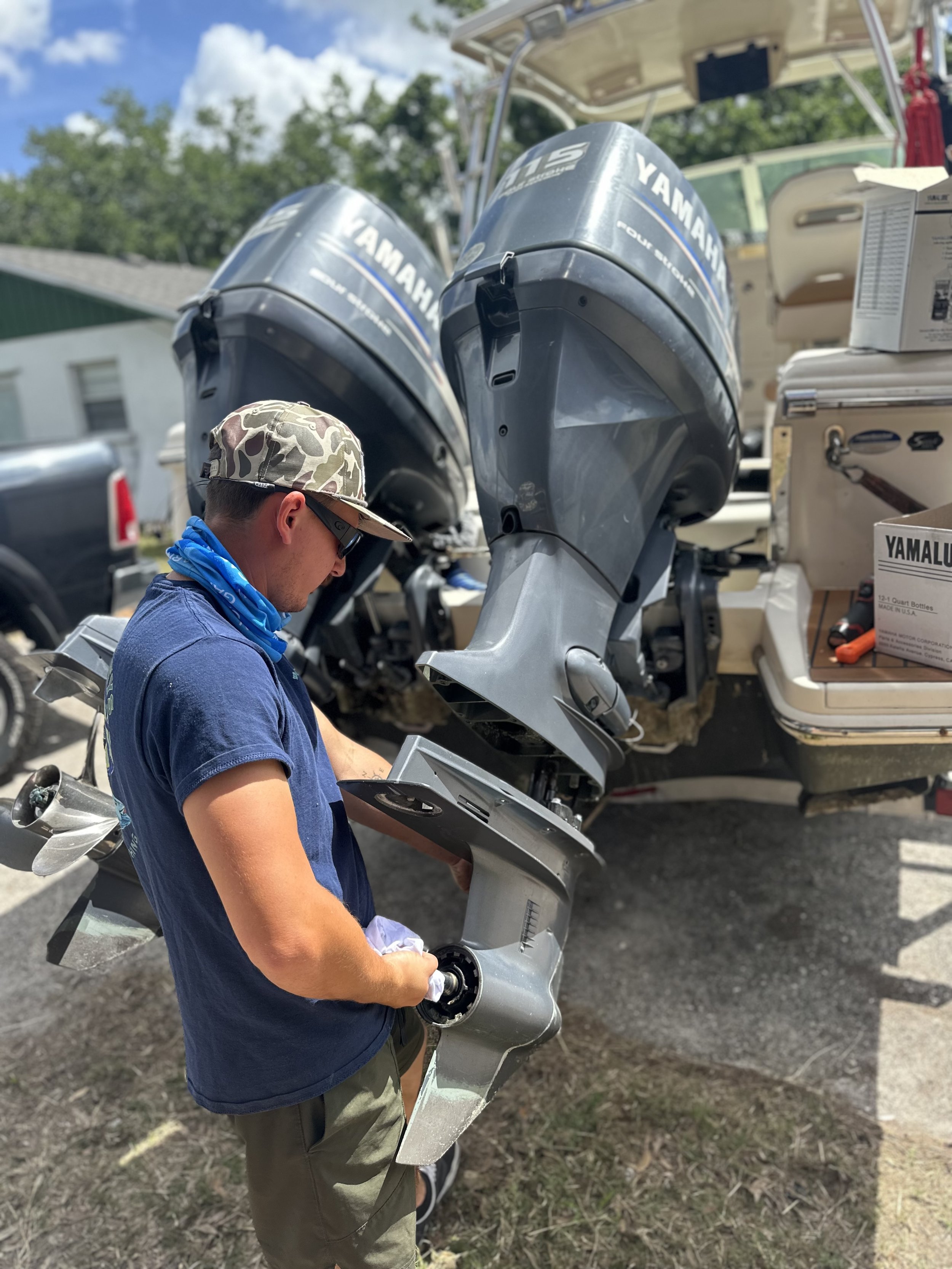 A young man wearing camouflage hat, sunglasses, and a blue bandana around his neck working on a silver outboard motor of a boat, with a white trailer or vehicle in the background and grassy ground.