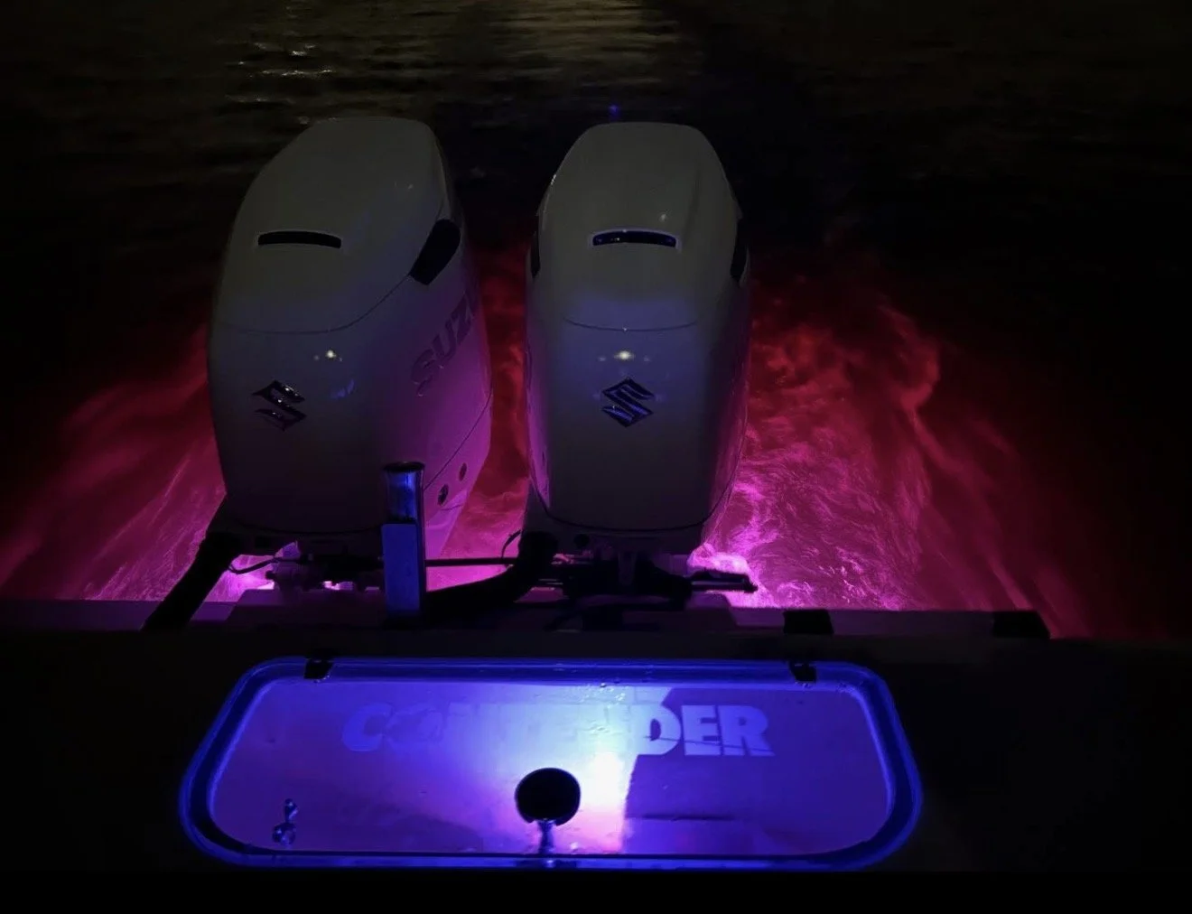 Two Suzuki outboard motors attached to the back of a boat, illuminated by blue and pink lights, with water visible behind them.