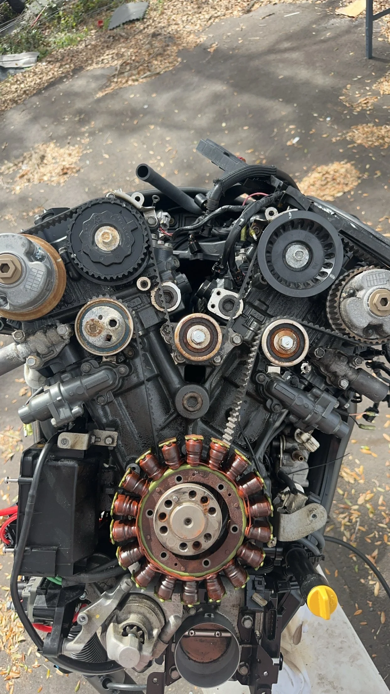 View of a disassembled motorcycle engine with visible gears, pulleys, and coils, outdoors on a paved surface with fallen leaves scattered around.