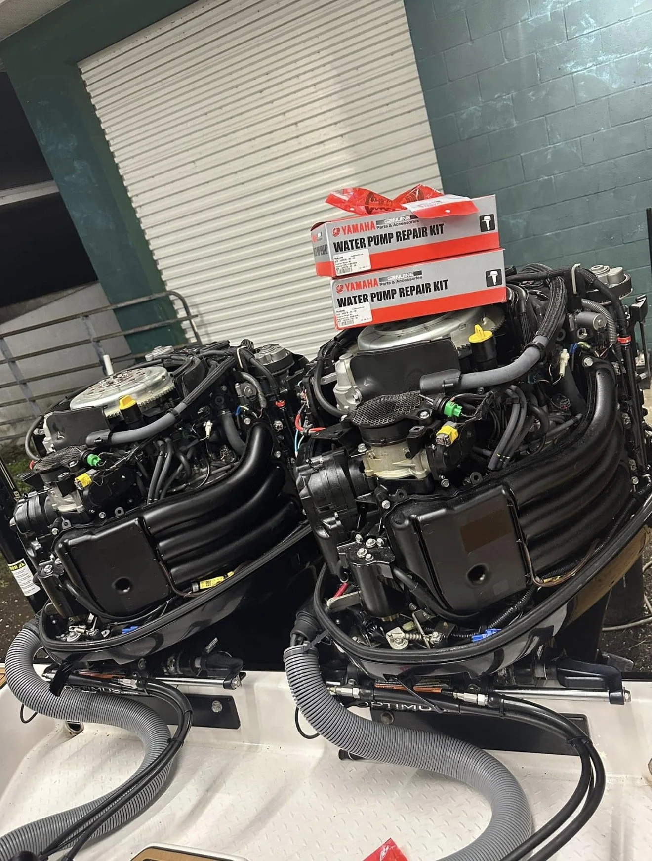 Two outboard motors with black and gray components, sitting on a white surface with gray hoses and wires, with boxes labeled Yamaha Water Pump Repair Kit in the background.