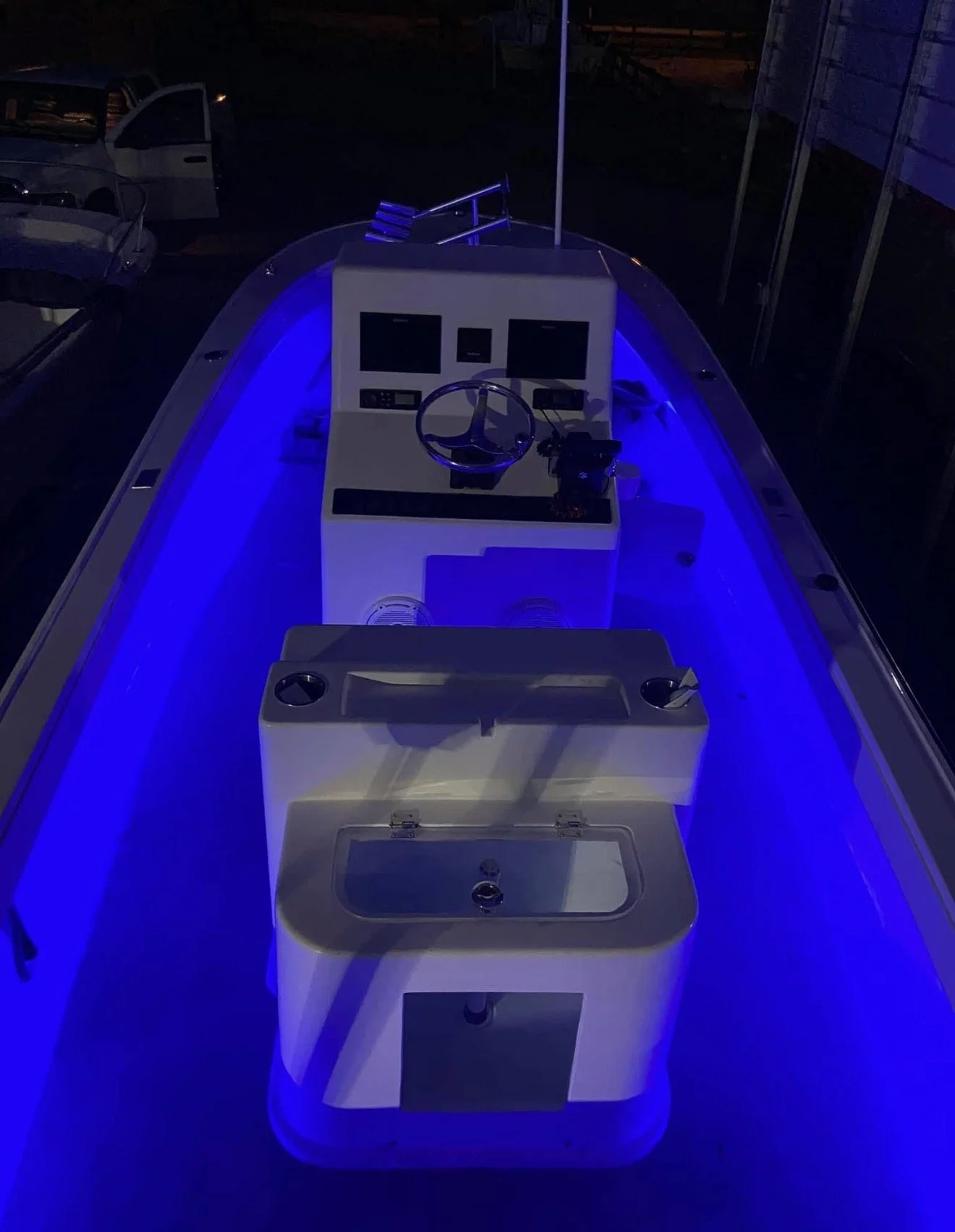 A boat with blue LED lighting along the edges, featuring a control console with multiple screens, a steering wheel, and a sink, docked at night with other boats visible in the background.