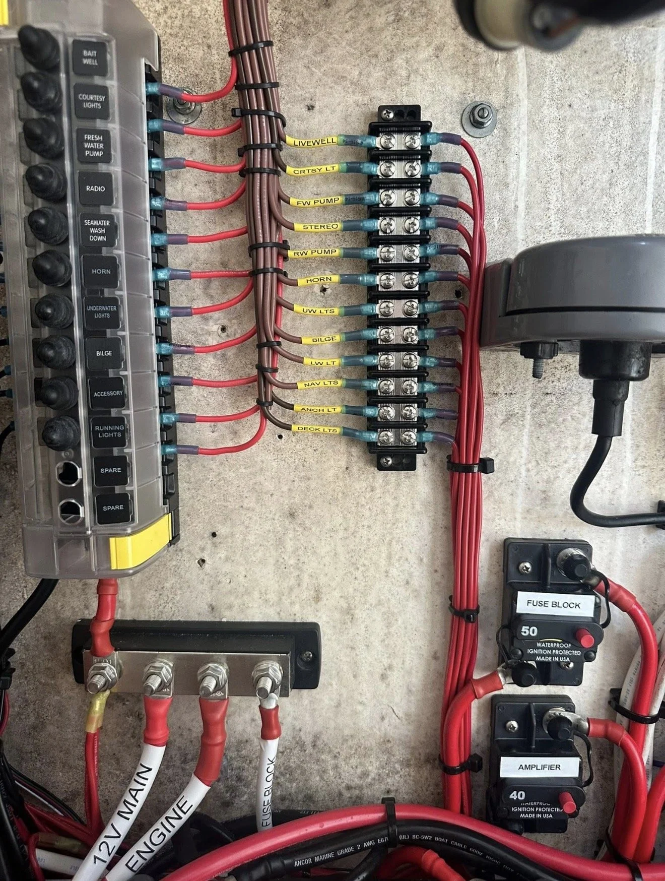A fuse box with multiple red and brown wires connected to various labeled terminals and circuit breakers. The labels include LIVEWELL, CRTSY LT, FW PUMP, STEREO, RW PUMP, HORN, UW LTS, BILGE, LW LTS, NAV LTS, ANCH LT, and DECK LTS. Additional components include a fuse block and an amplifier, with wires labeled MAIN, ENGINE, and FUSEBLOCK.