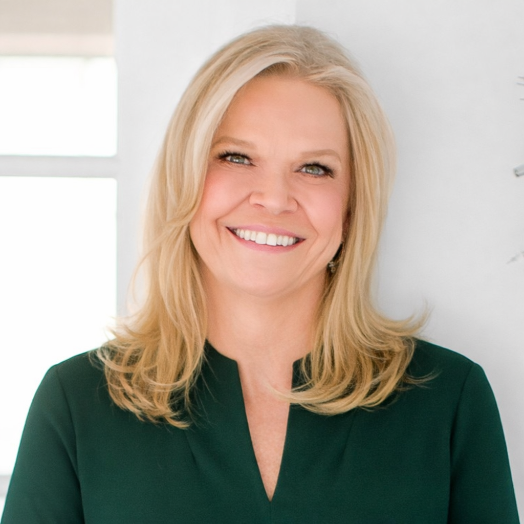 A smiling blonde woman in a dark green top in an office setting.