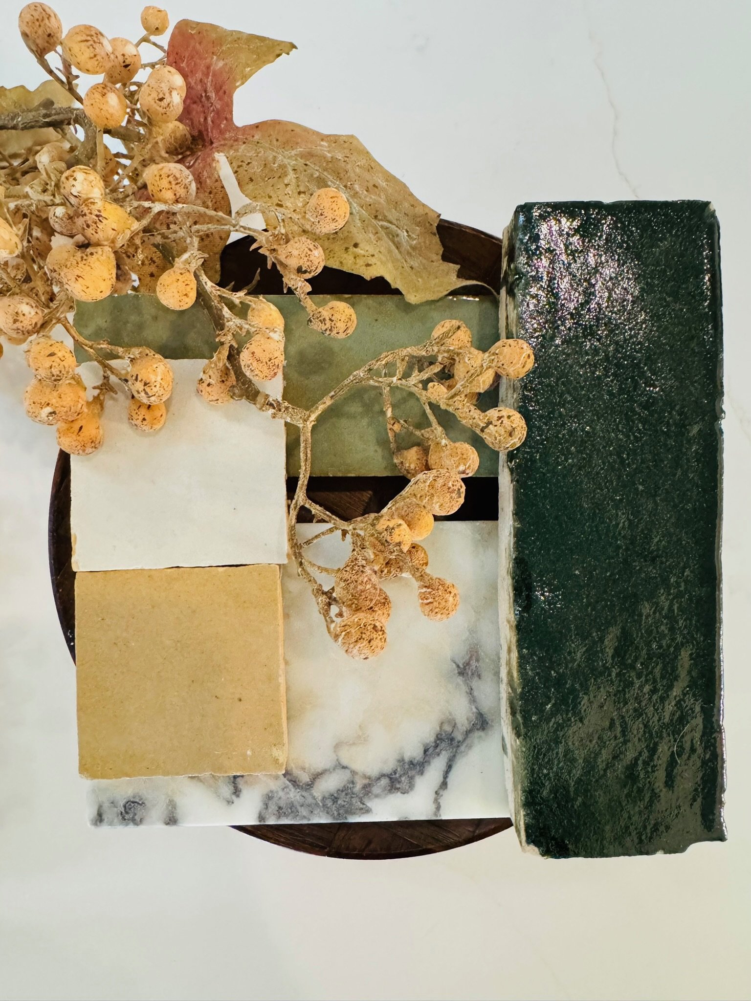 A collection of quatrefoil-shaped soaps, including a dark green bar and several beige and white bars, arranged with dried flowers and leaves on a white marble surface.