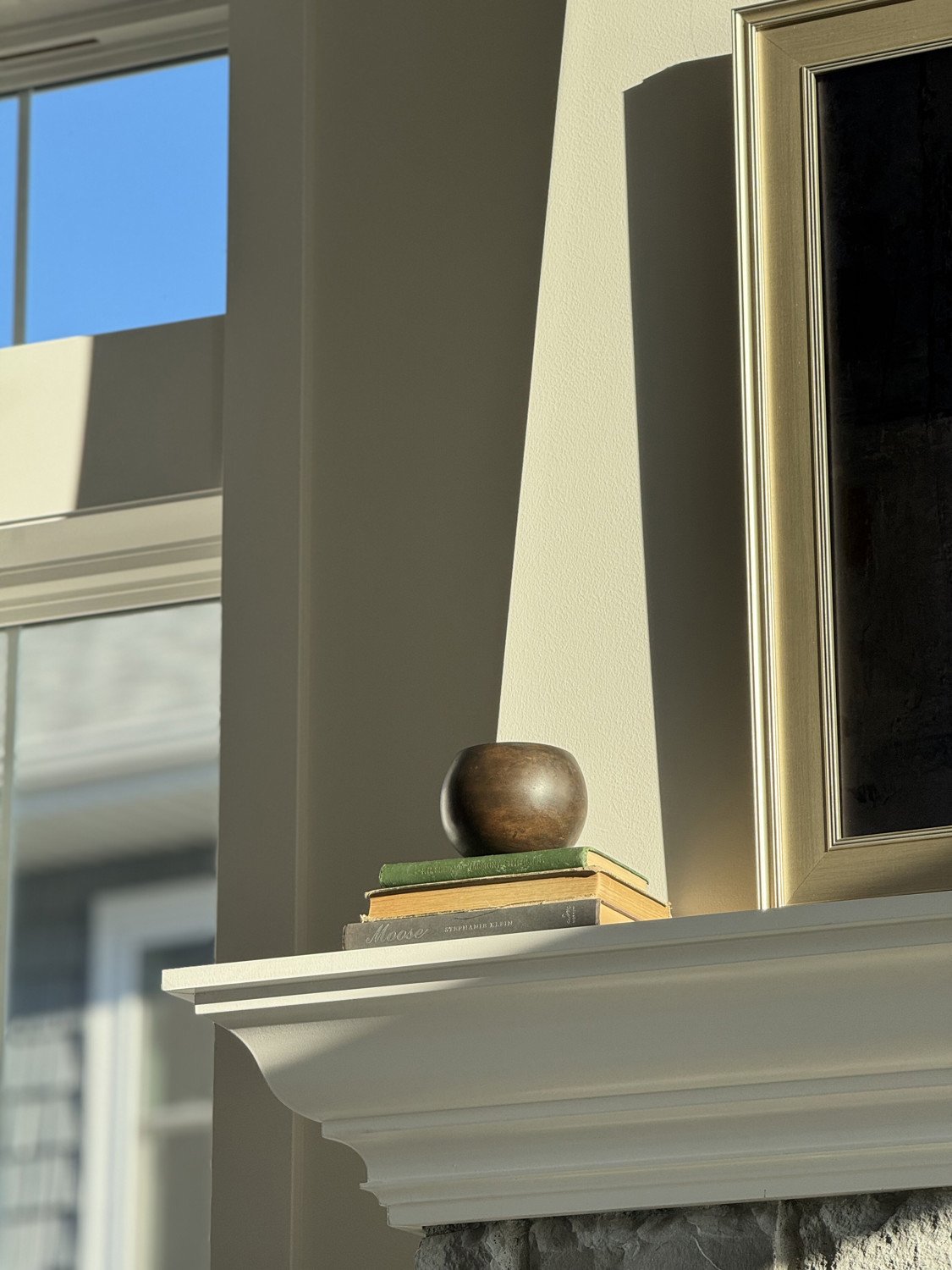 A wooden shelf holding a small brown bowl on three stacked books, part of a fireplace mantle in a sunny room with windows showing the blue sky outside.