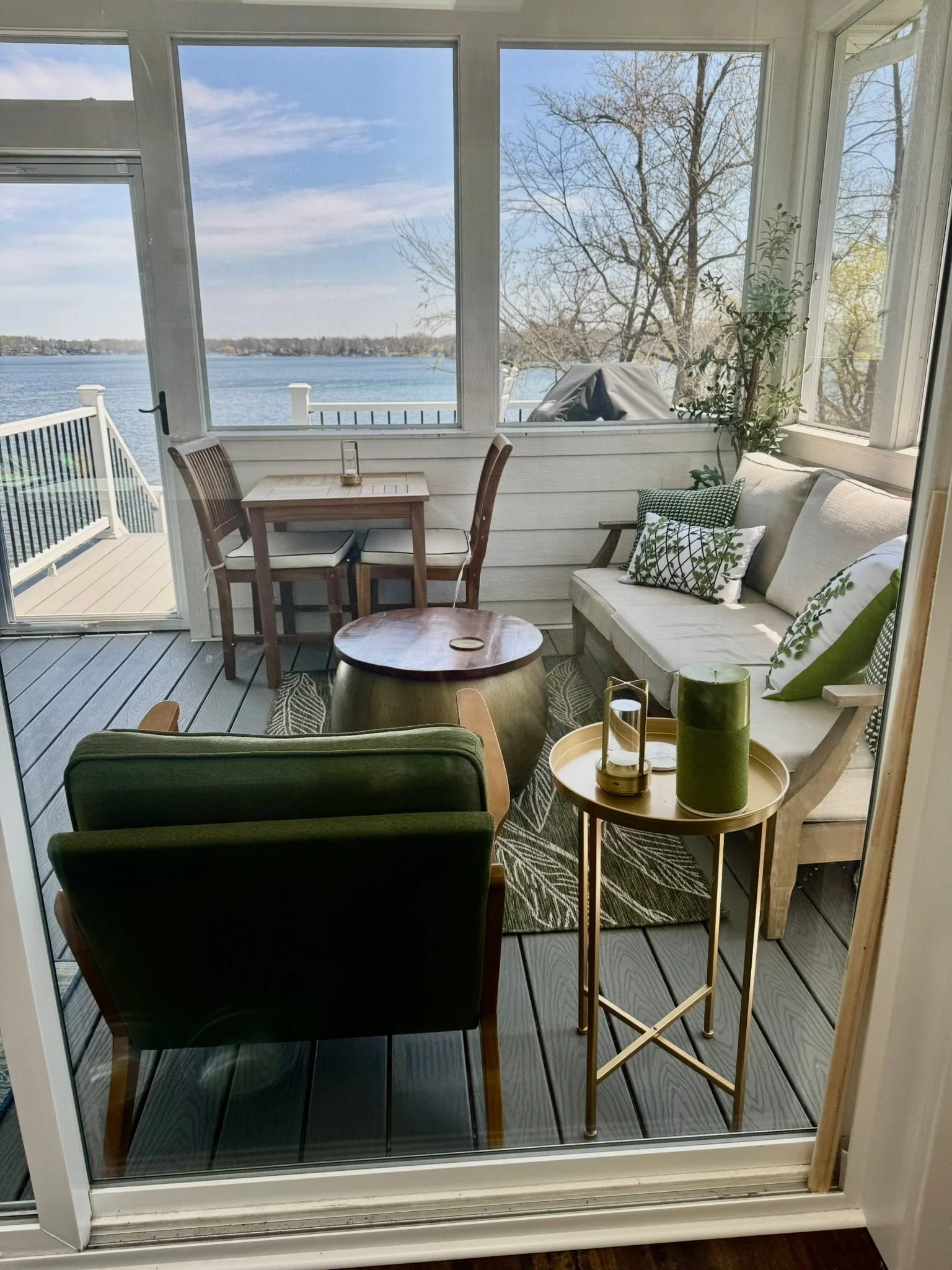 View of a screened porch overlooking a lake, with outdoor furniture including a table with two chairs, a cushioned sofa with pillows, and decorative items on a side table, trees in the background.