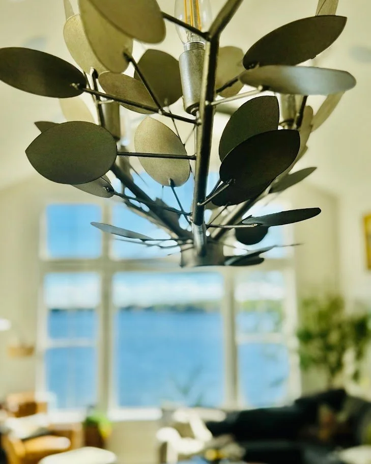 Close-up of a modern chandelier with leaf-shaped elements hanging from it, with a bright room and large window overlooking a body of water in the background.