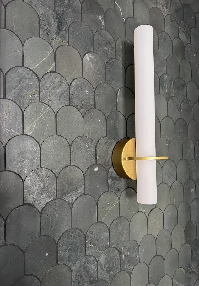 Modern wall sconce with a white cylindrical shade and gold base mounted on gray tile wall with scalloped pattern.