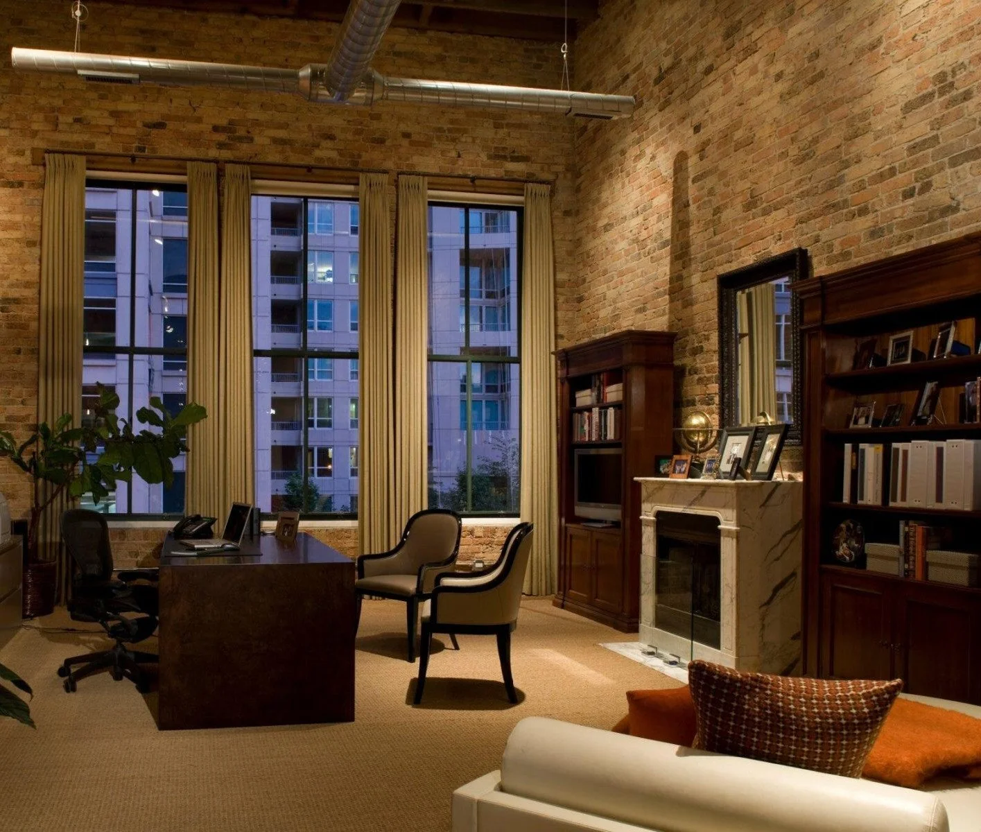 Modern office with brick walls, large windows with beige curtains, a wooden desk with an office chair, beige armchairs, a fireplace with a marble mantel, bookshelves, and framed photos on top of the fireplace and shelves.