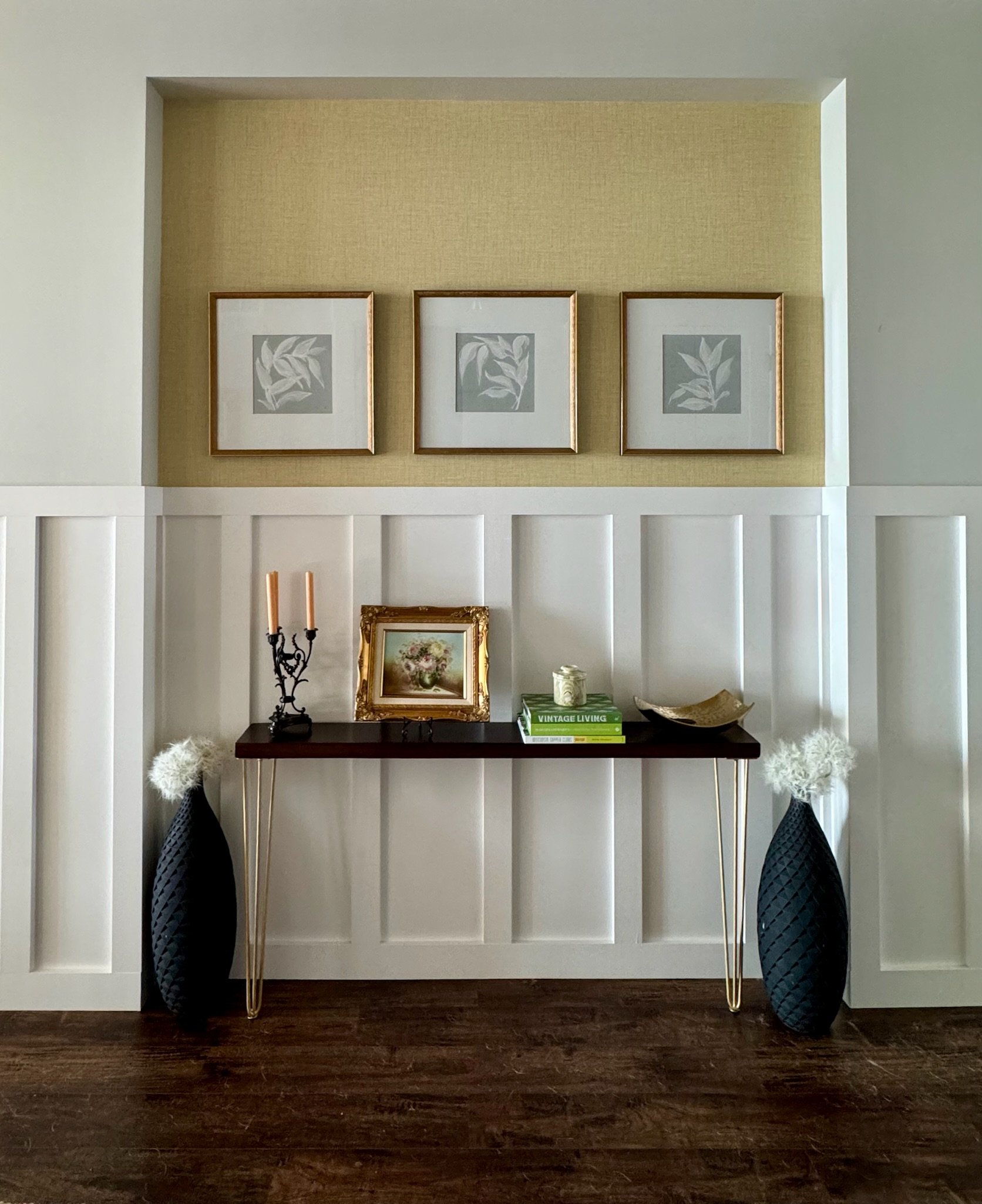 Interior living space with built-in wall niche, three framed botanical prints, a dark wood console table with framed artwork, books, a small plant, and decorative objects, flanked by tall black textured vases with white pampas grass, and dark hardwood flooring.