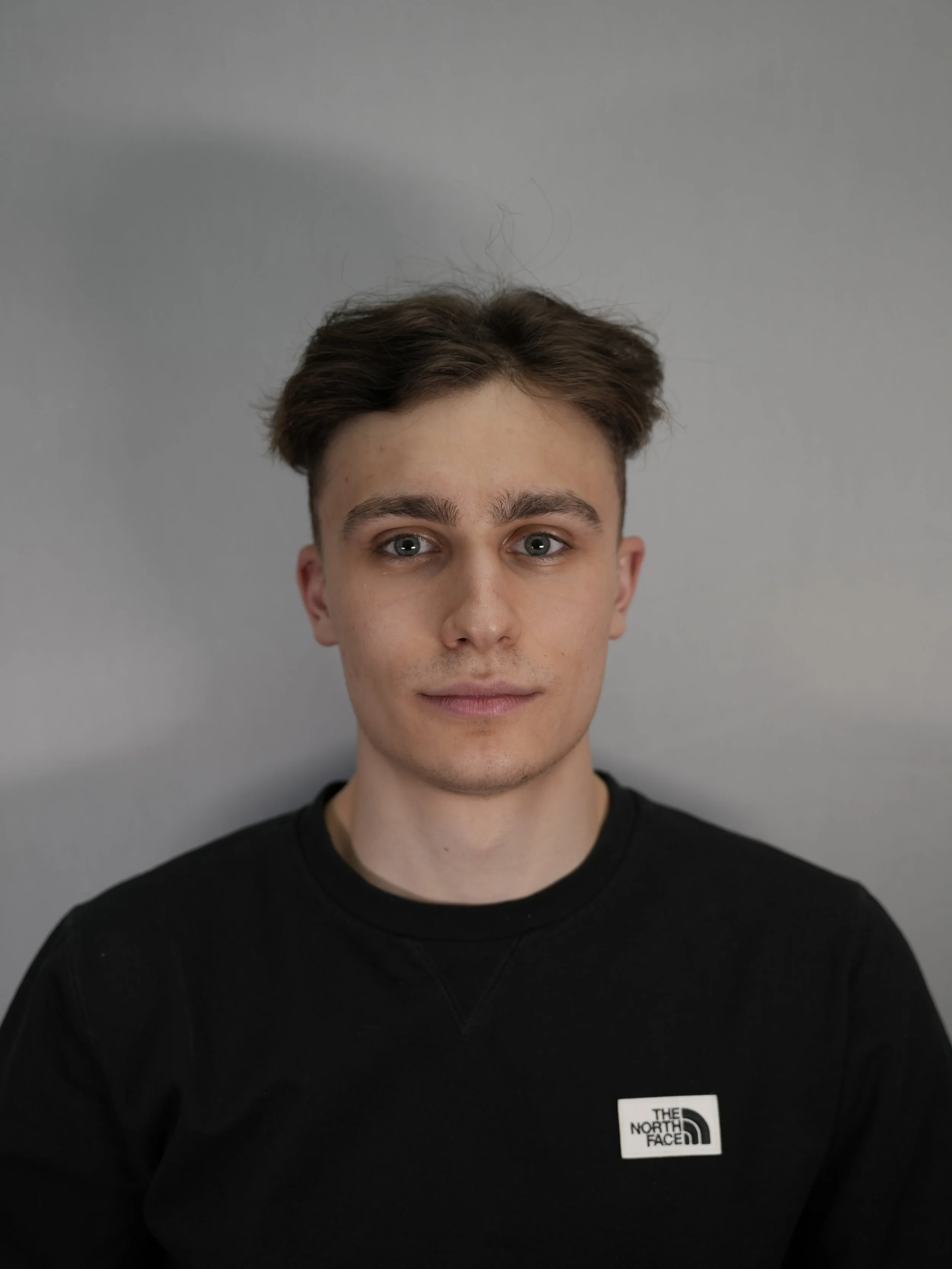 A young man with short brown hair and blue eyes wearing a black The North Face sweatshirt, standing against a plain gray background.